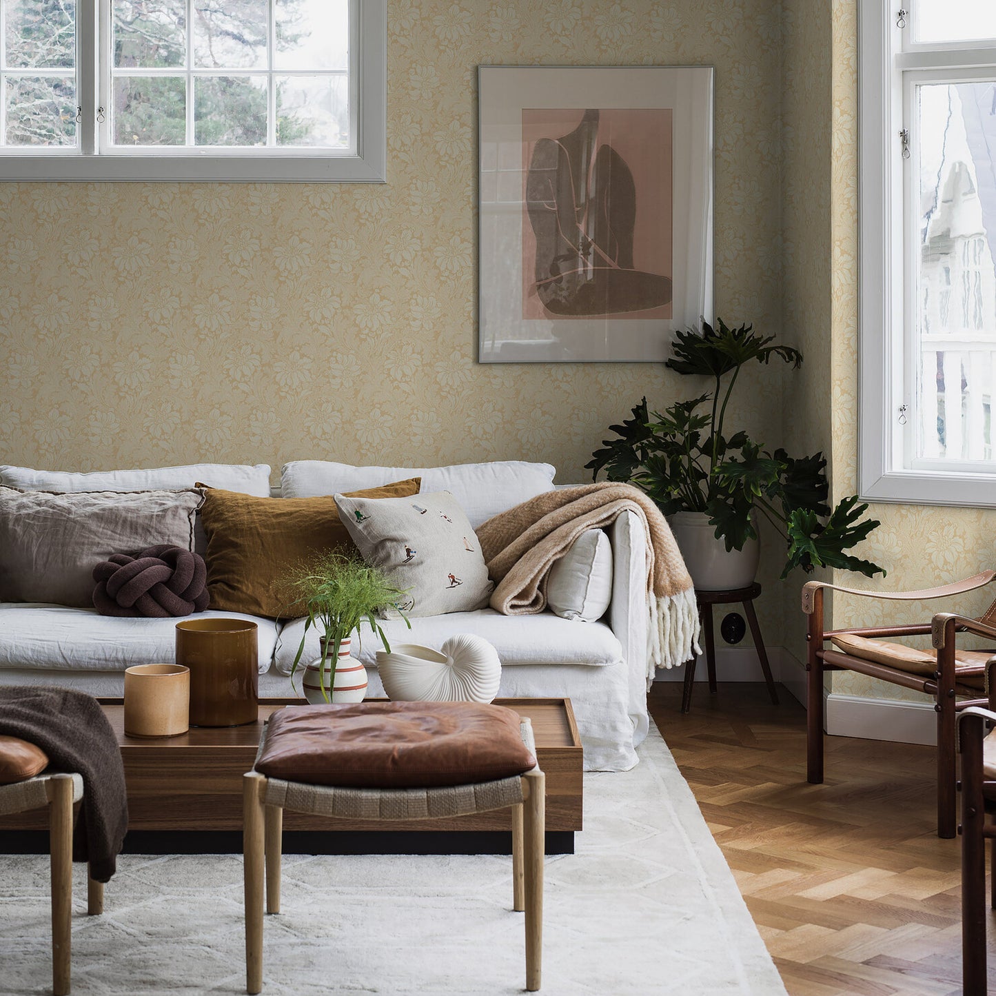 Living room featuring a wall covered with yellow botanical wallpaper, light gray sofa, cushions, wooden coffee table, leather ottoman, plants & abstract art.