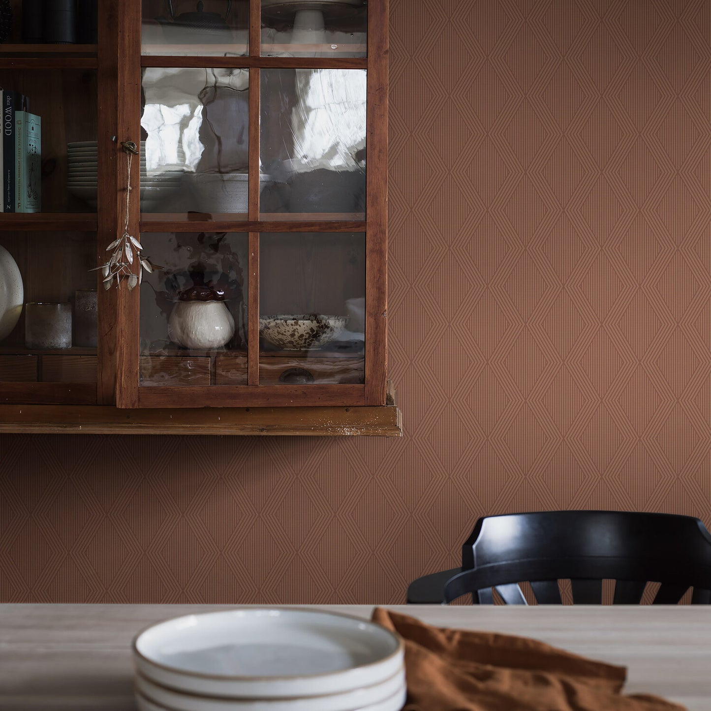 Terracotta & gold wallpaper on a brown textured wall, with a wooden cabinet with glass doors and a table set with white plates, a brown napkin, and a black chair.