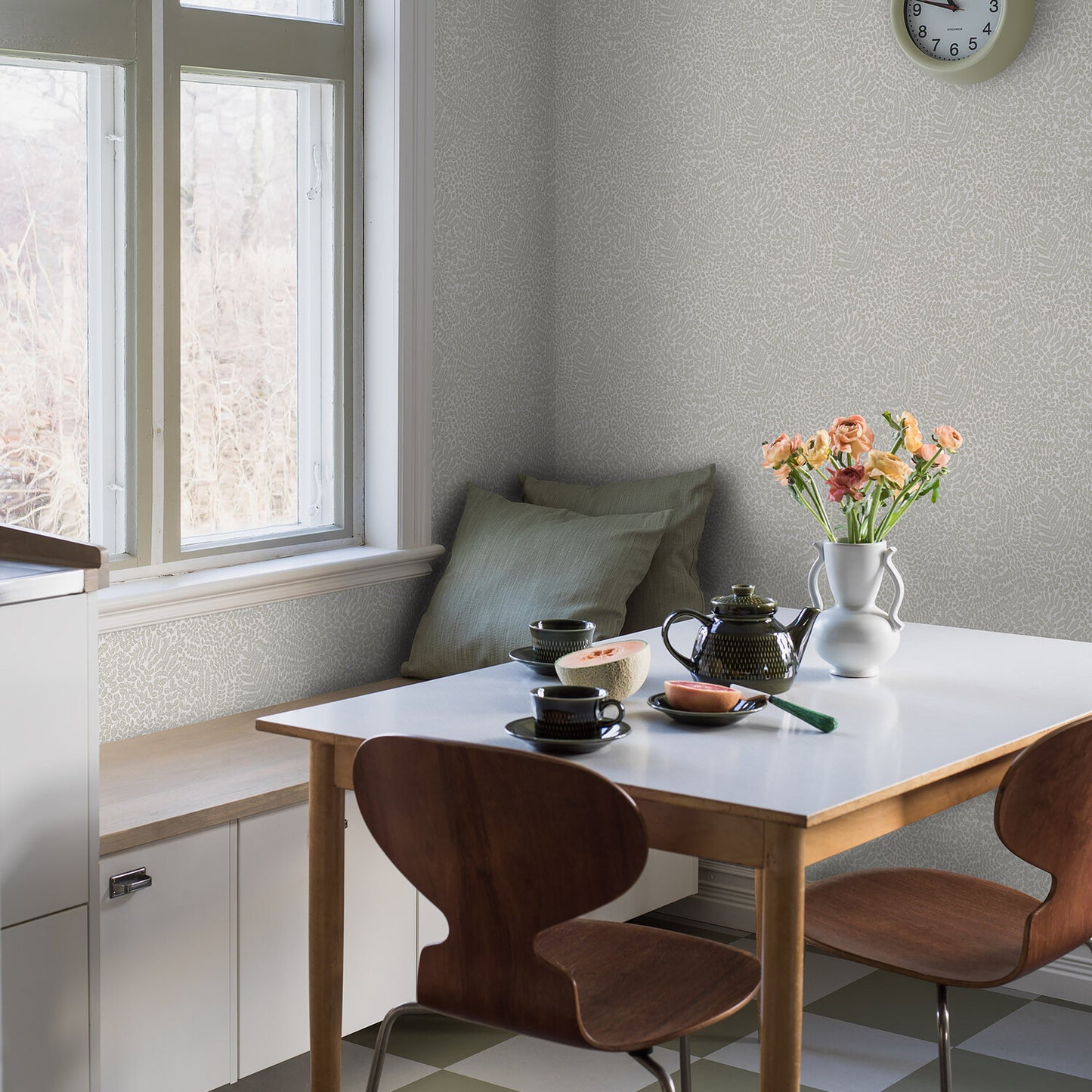 Wallpaper with sage tones on the wall behind a window-side dining nook; white table, wooden chairs, bench cushions, teapot, cups, melon half, and a vase of peach flowers.