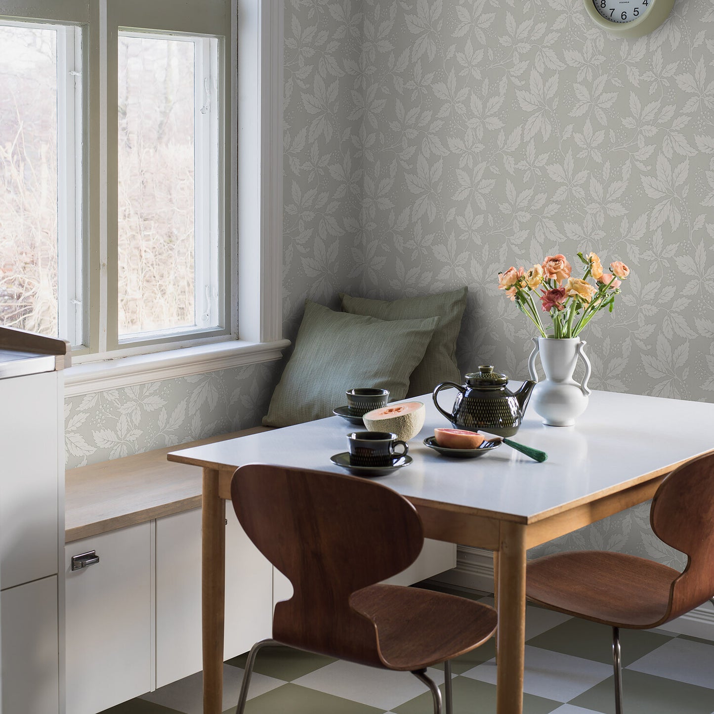 Bright kitchen nook with white table, wooden chairs, bench, leaf-pattern wallpaper, vase of orange flowers, teapot and cups.