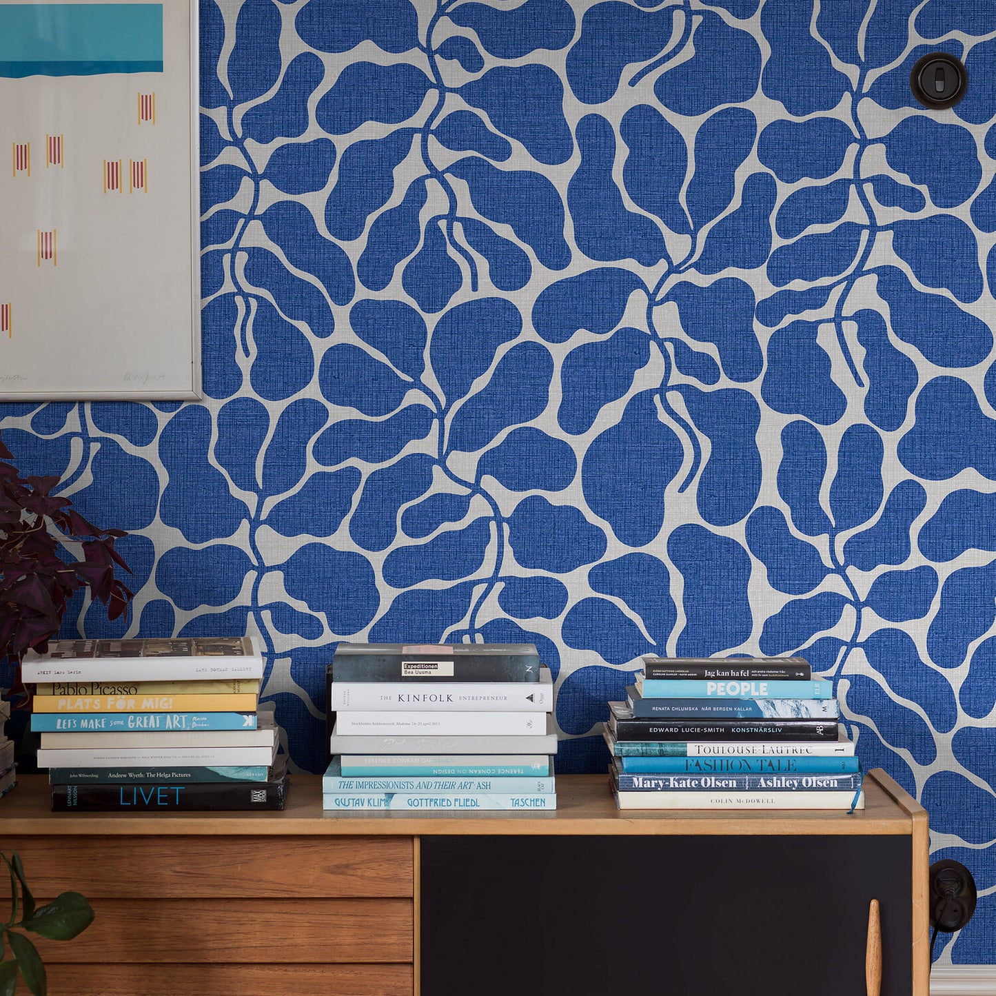 Bold blue leaf-pattern wallpaper behind a wooden sideboard with stacked books and a potted plant
