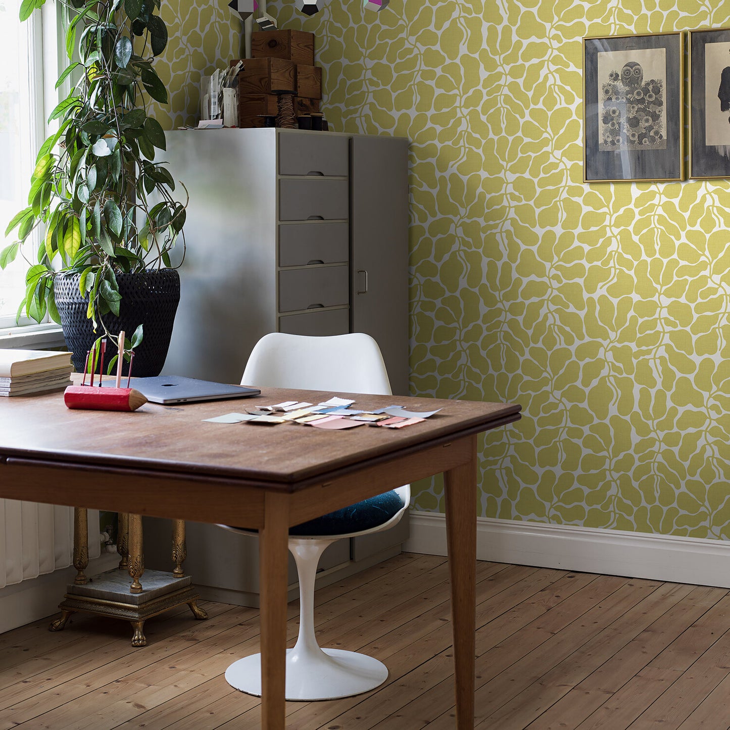 Wooden desk with white tulip chair in lime-patterned office, plant, gray filing cabinet, scattered papers and a laptop.