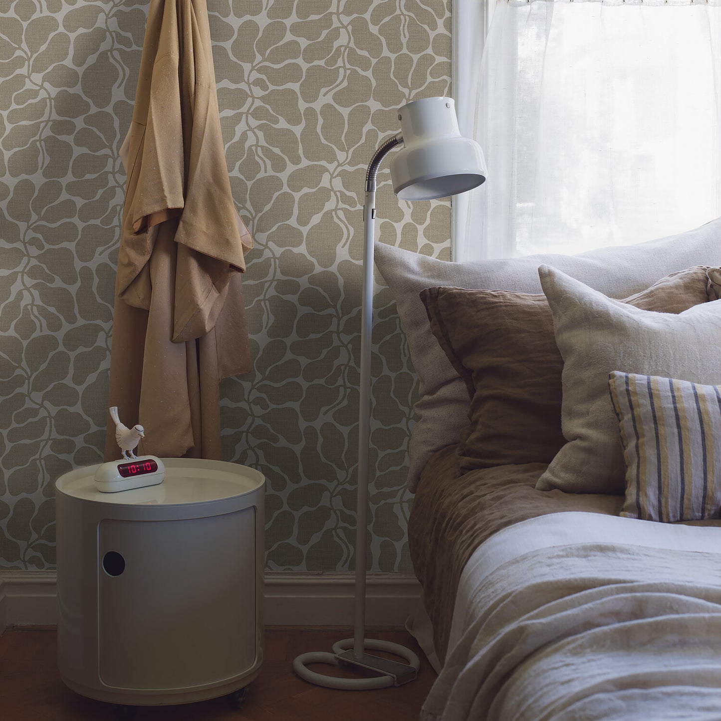 Cozy bedroom corner with beige robe, round light-gray bedside table, tissue box, white floor lamp, and bed with brown/cream pillows.