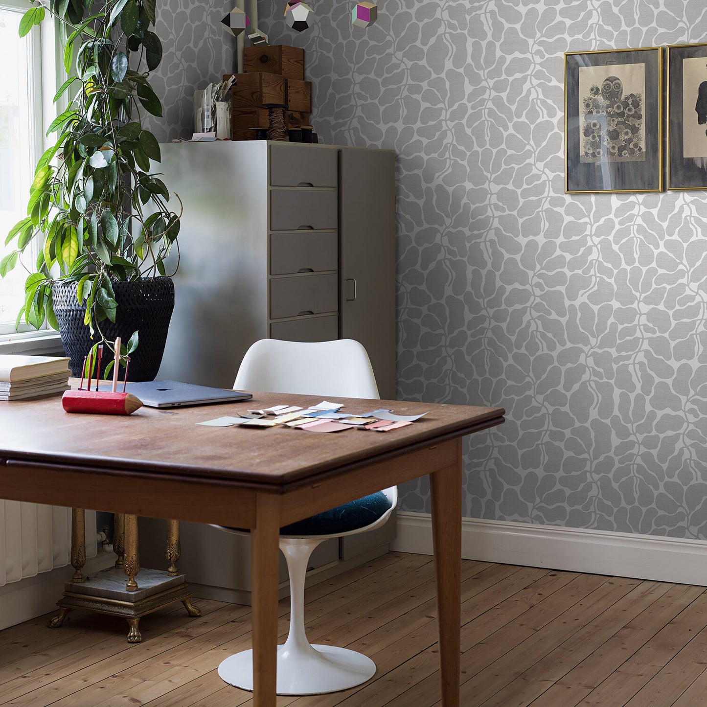 Wooden desk with white tulip chair in a stylish grey patterned wallpaper office; tall plant, filing cabinet, scattered papers.