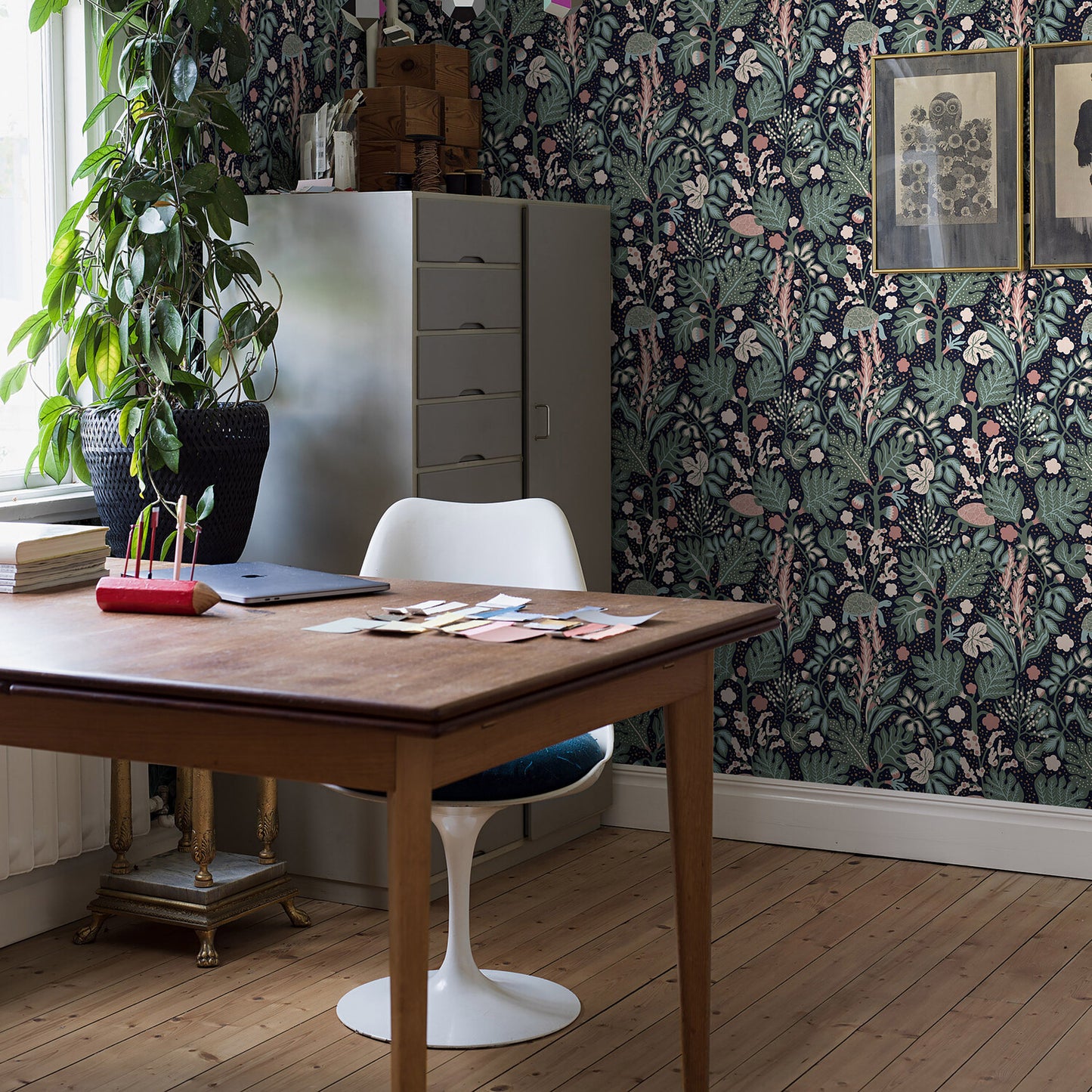 Wooden desk with white Tulip chair in a vintage home office; leafy wallpaper, tall filing cabinet, potted plant.