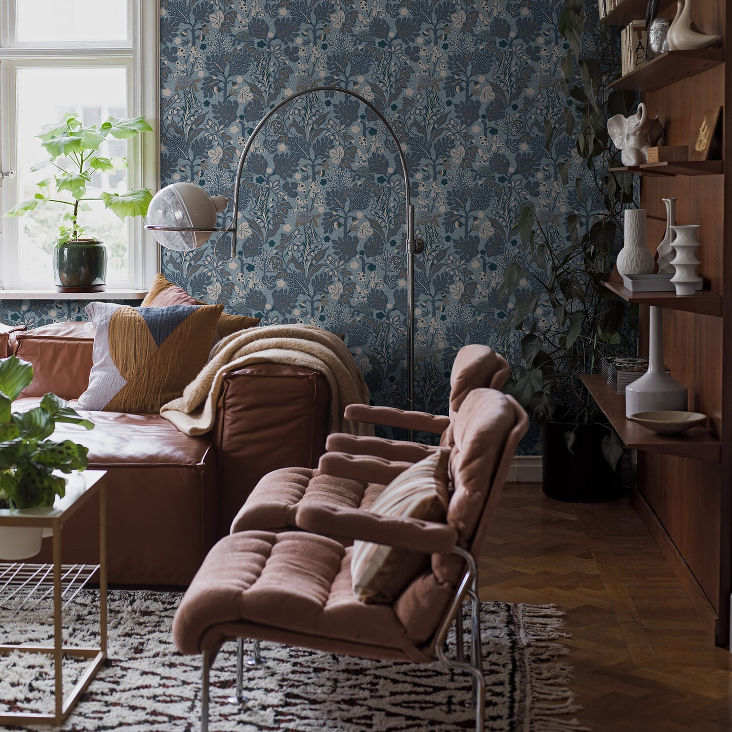 Tan leather sofa with tufted lounge chair, arched floor lamp, blue floral wallpaper, wooden shelves and plants.