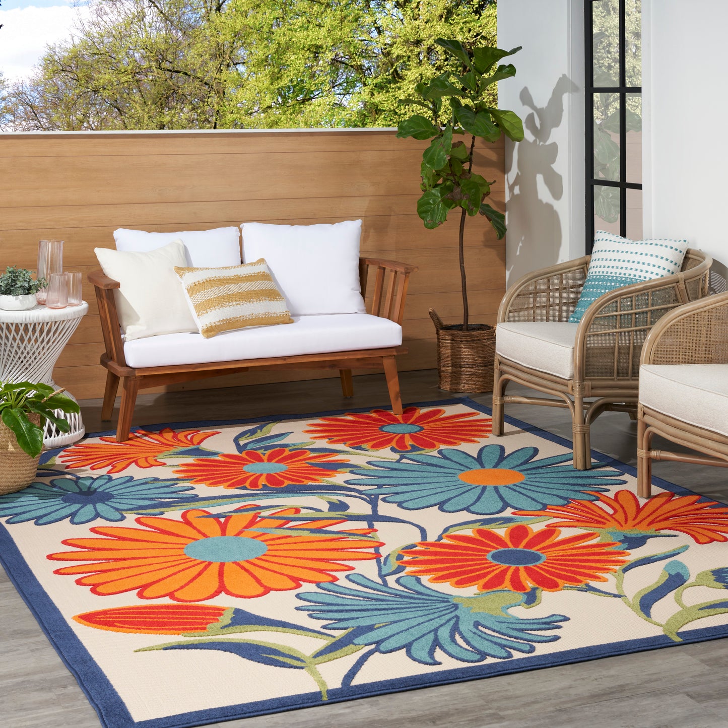 Outdoor seating with a bright orange and blue floral rug, white-cushioned wooden sofa, wicker chairs, and potted plants.