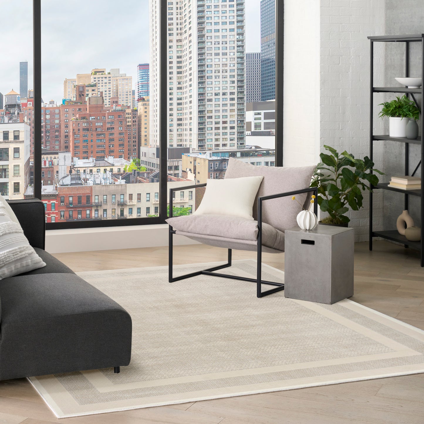 Modern living room with city view, beige rug, gray sofa, metal-framed chair, concrete side table and greenery.