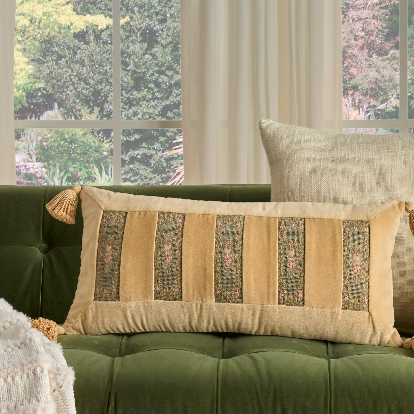 Green velvet tufted sofa with a beige striped rectangular lumbar pillow, floral panel accents, and tassel ends.