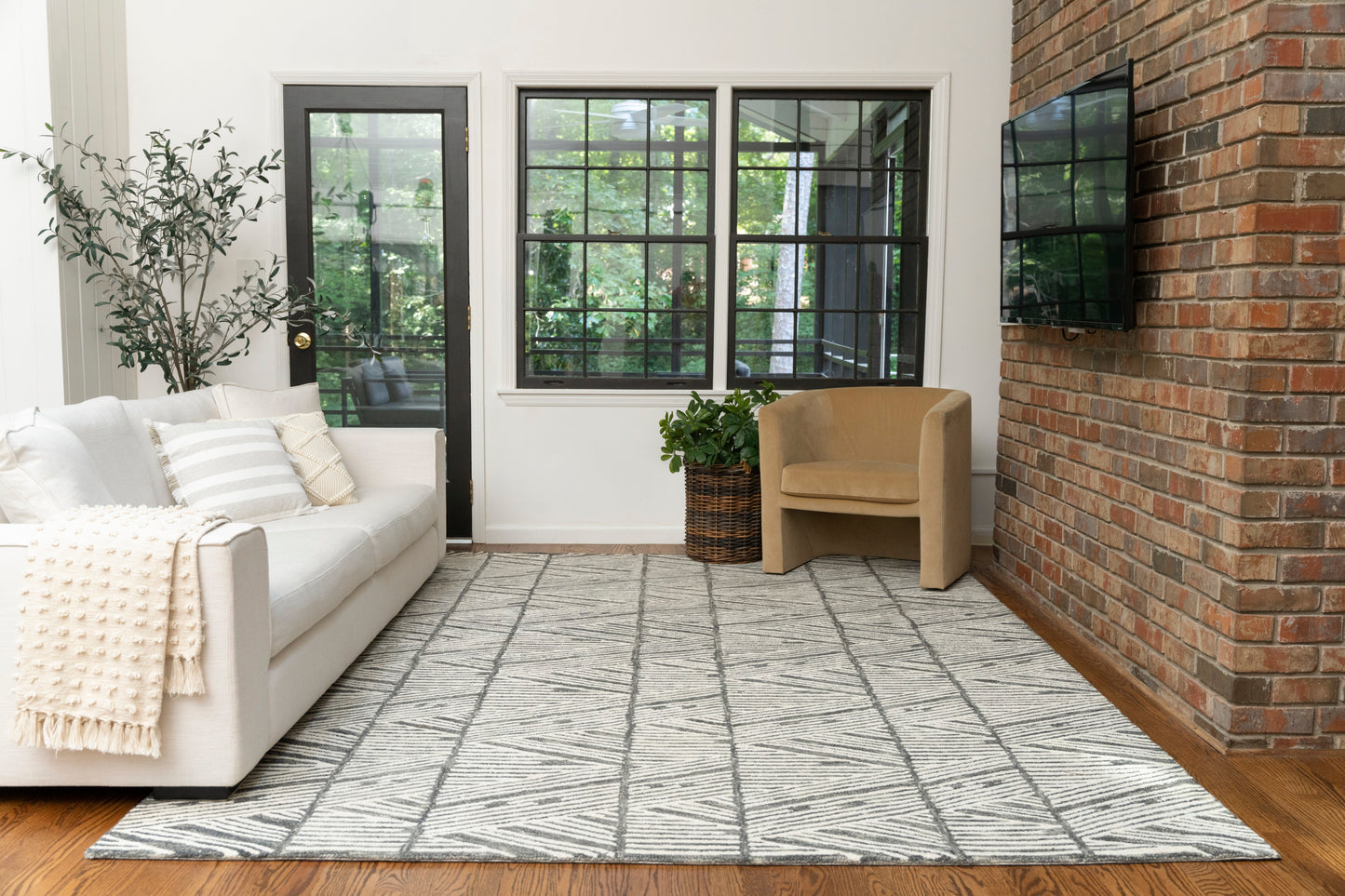 Gray rug in a modern living room scene; 8' x 10' size; cream sofa with a beige throw, beige armchair, brick wall, flat-screen TV, potted plant, and black-framed windows.