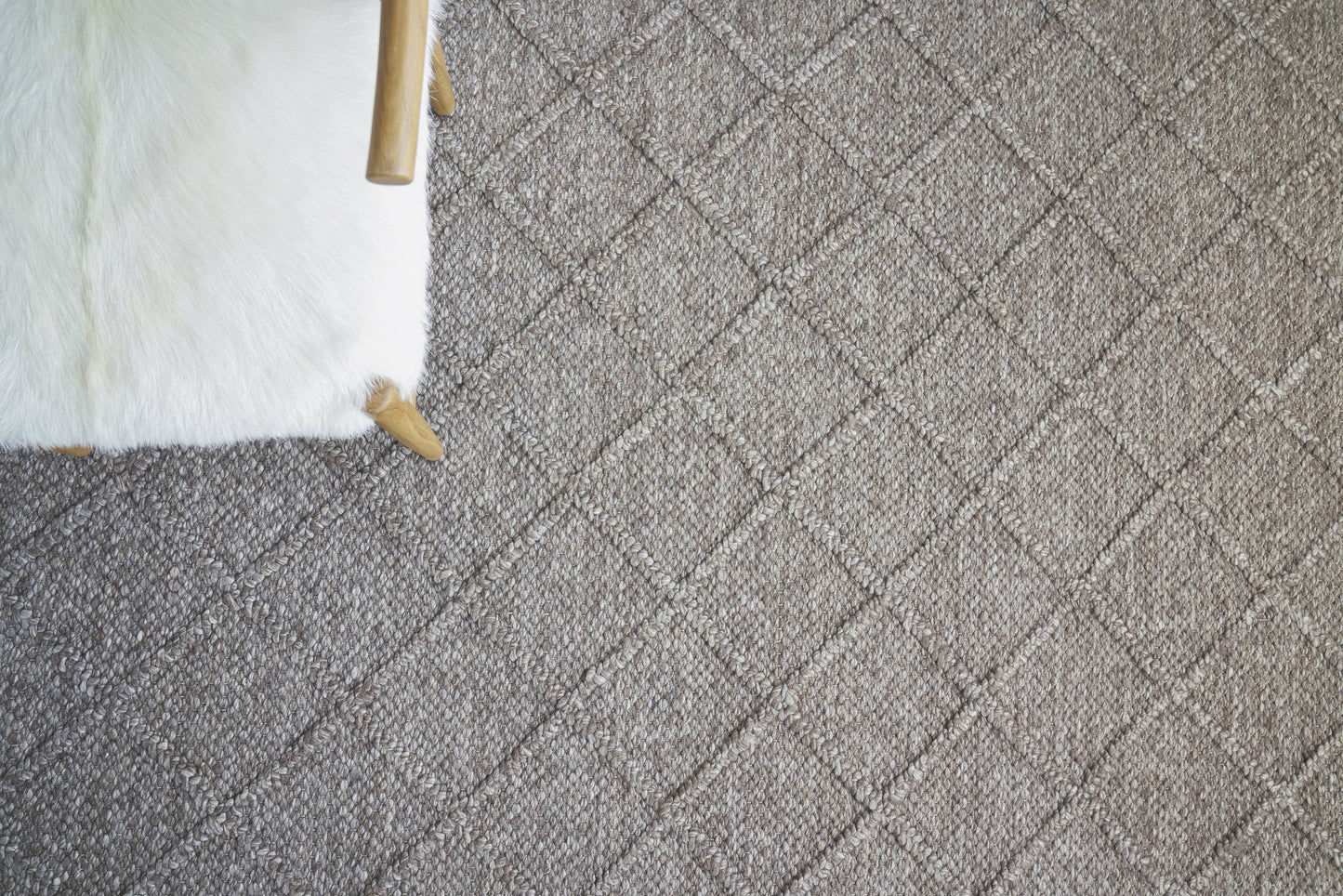 Beige textured rug with a diamond lattice pattern; white fluffy ottoman with light wood legs at the edge.