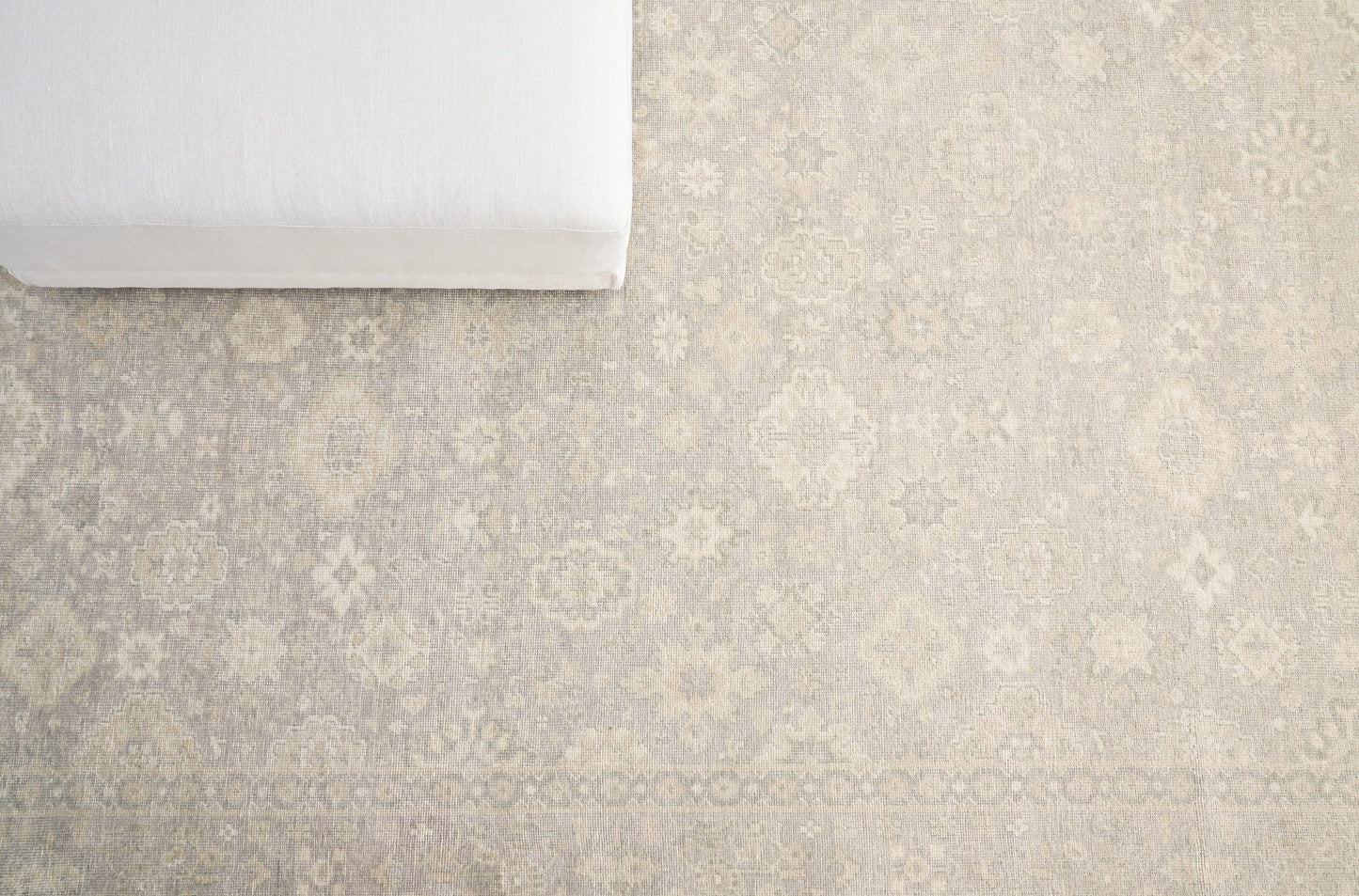 Beige rug with subtle damask motif in ivory; white cushion resting at the top-left corner.