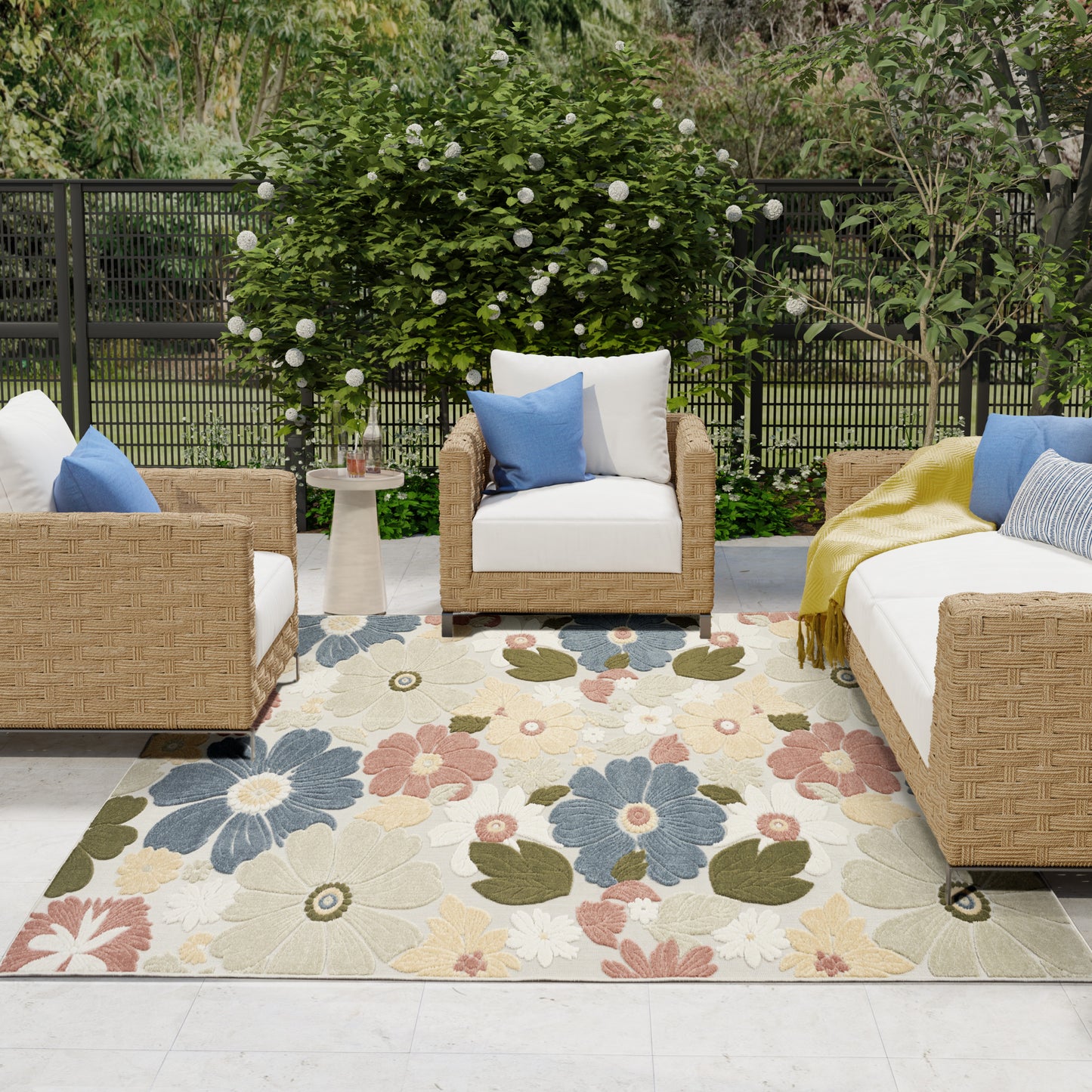 Outdoor wicker seating with white cushions and blue pillows on a pastel floral rug; garden backdrop.