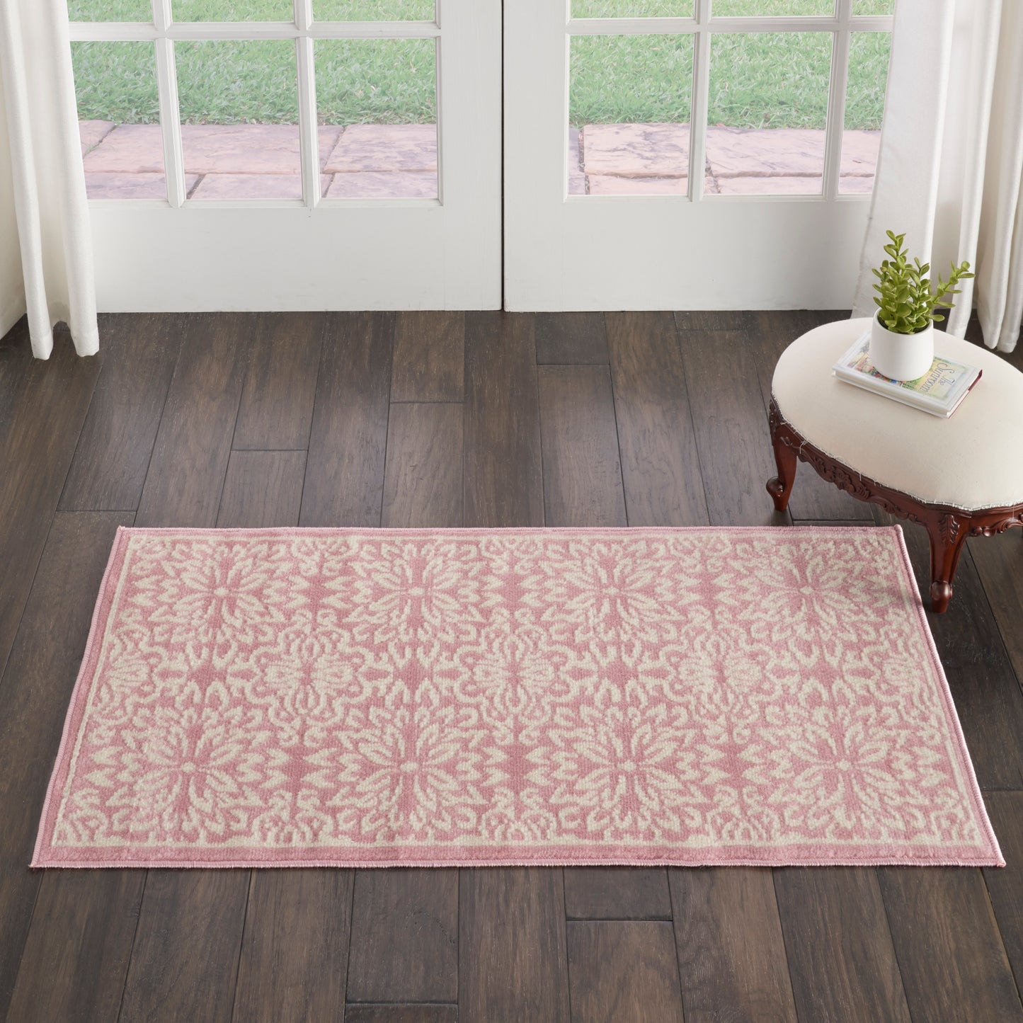 Rectangular pink rug with cream damask pattern on dark wood floor near French doors, beside a small side table with plant.