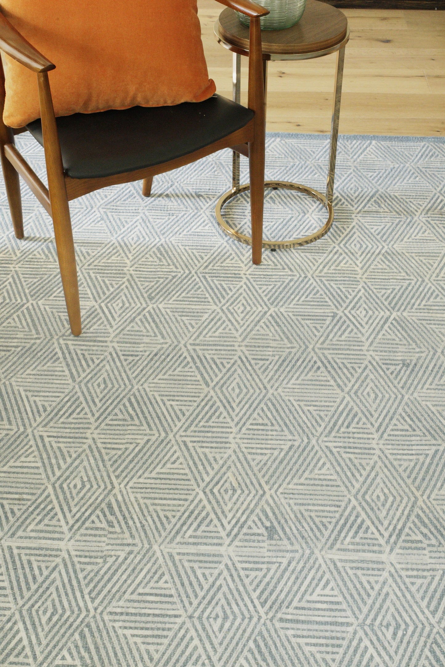 Geometric flat-weave rug on the floor with a mid-century wooden chair (black seat, orange cushion) and a round gold metal side table nearby.