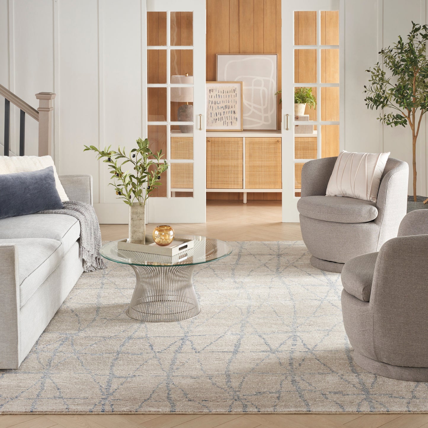 Light gray living room with round glass coffee table, gray sofas & armchairs, pale blue patterned rug, wooden doors.