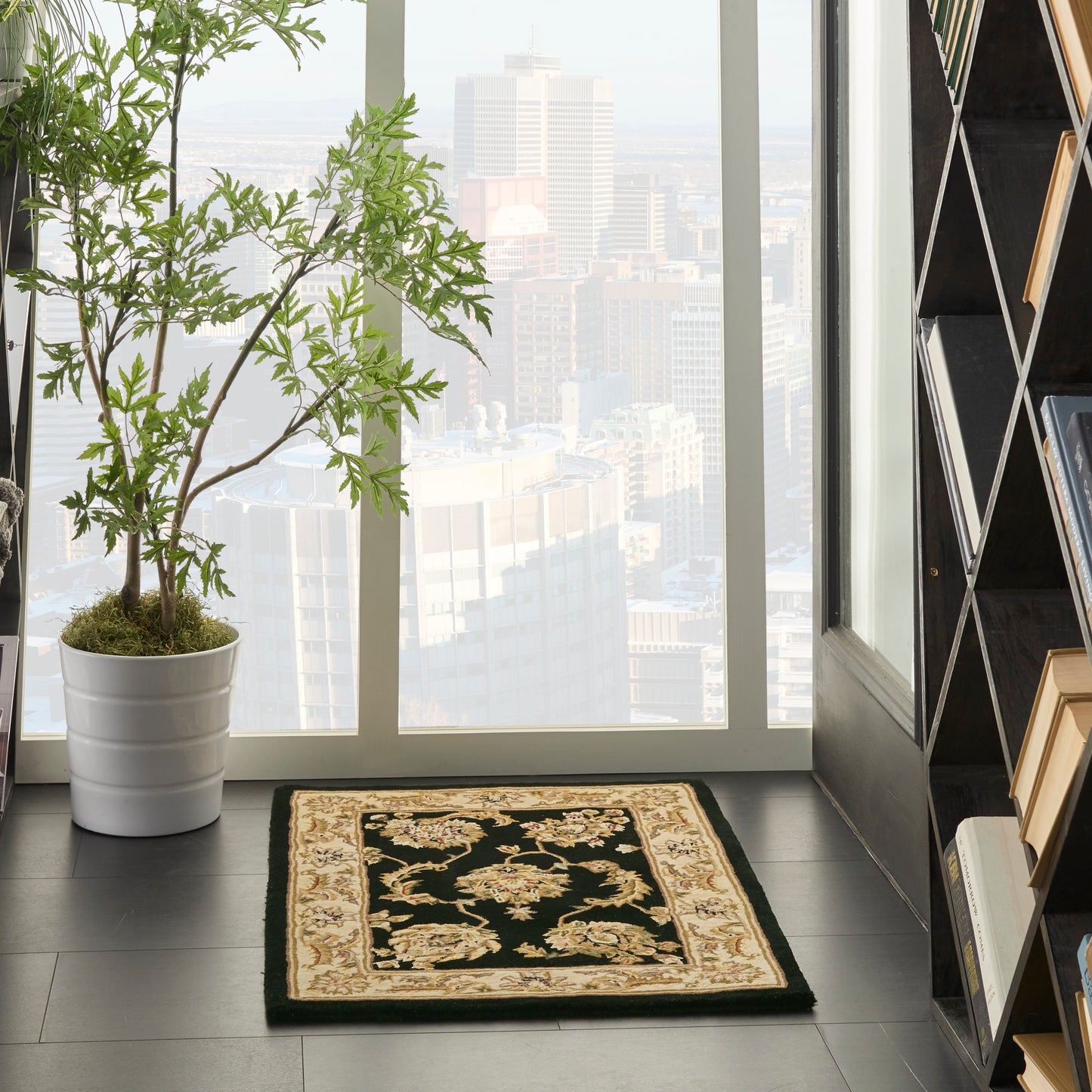 Ornate black area rug with gold/beige border on dark floor beside a potted plant in a bright urban loft.