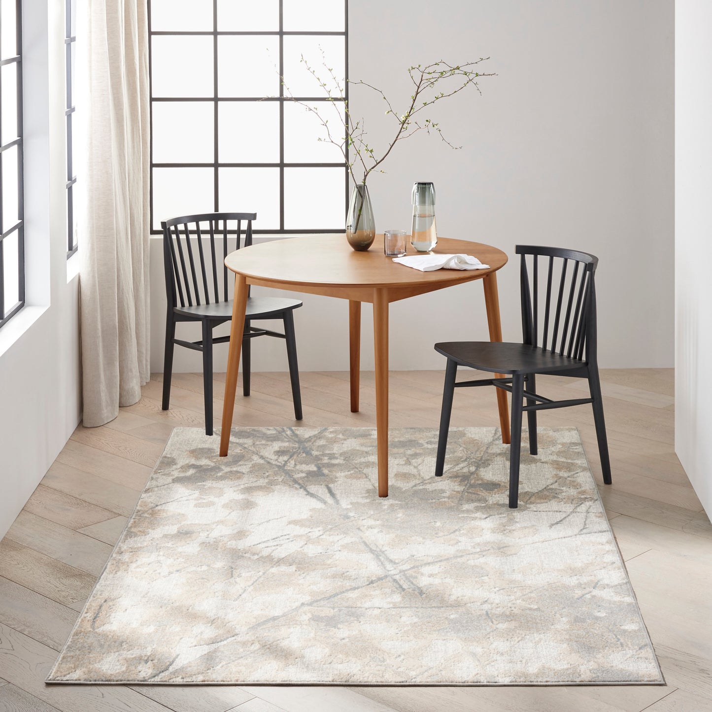 Light patterned rug beneath a round wooden dining table with two black chairs, a glass carafe, and a vase with branches by grid windows.
