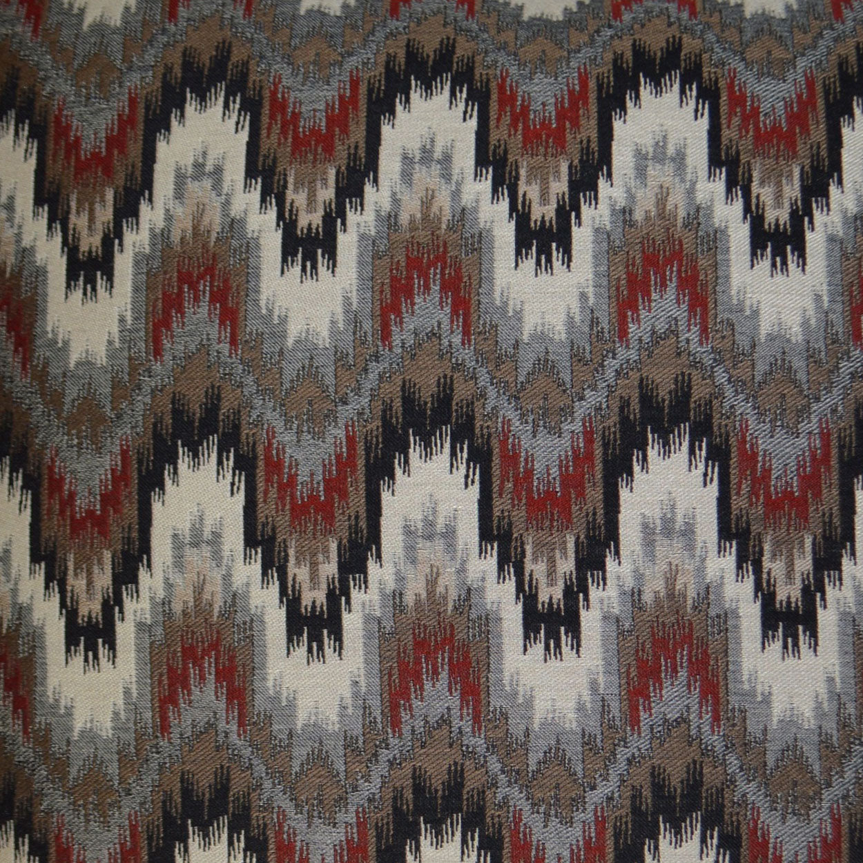 Multi-color zigzag ikat fabric in gray, charcoal, red & brown tones; woven chevron pattern for upholstery or decor.