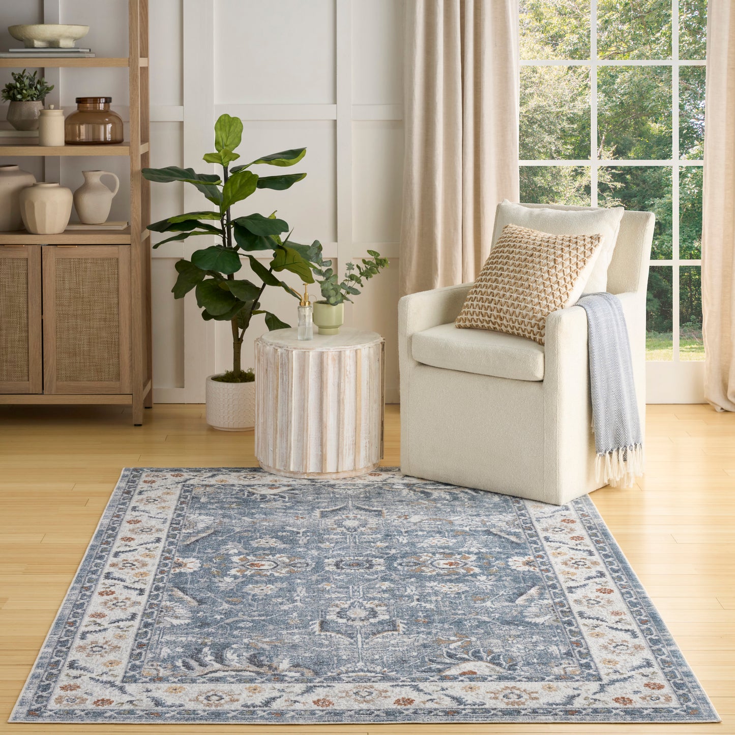 Blue and ivory traditional area rug in a bright living room with a cream armchair, throw, round ribbed side table, and plants.