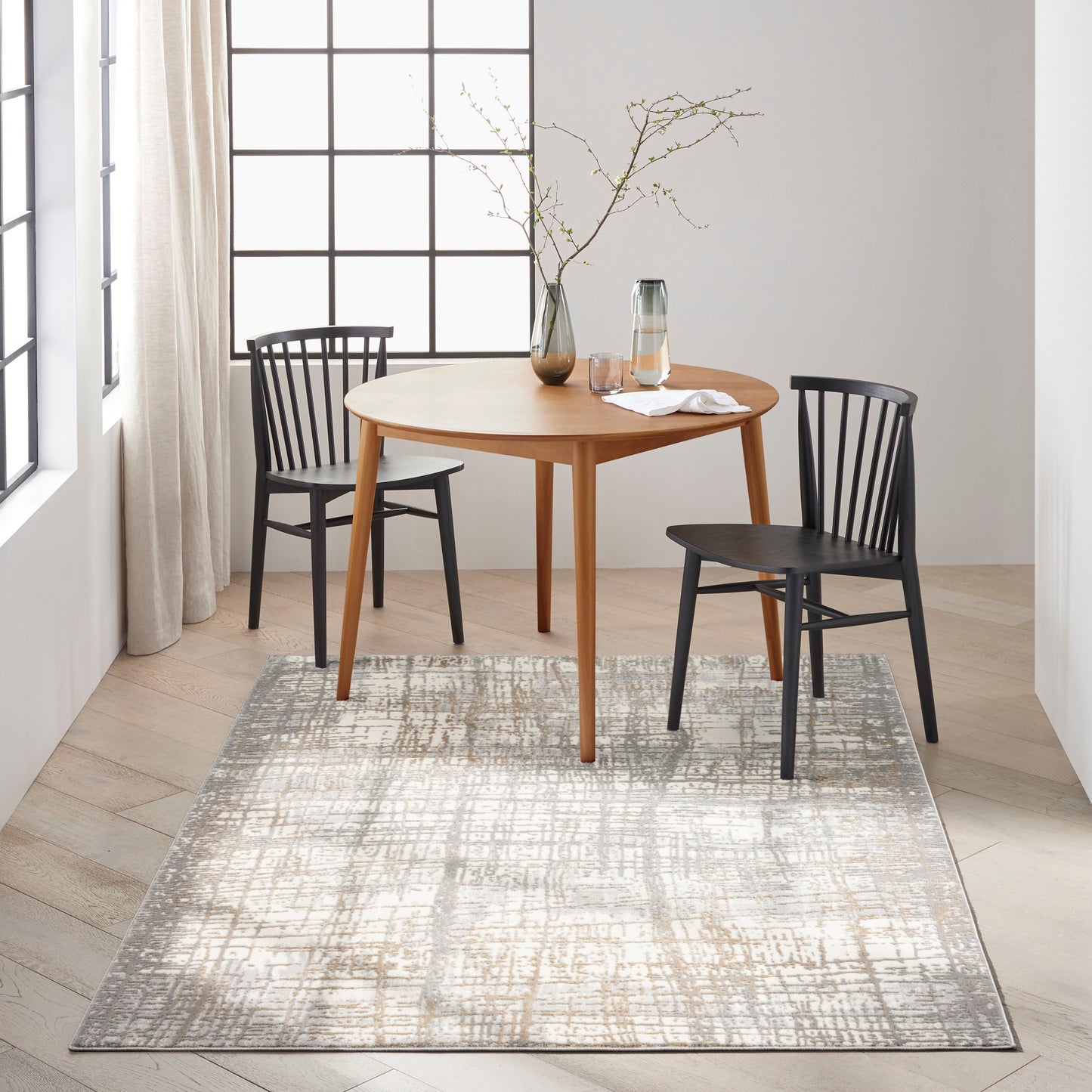 8' x 10' modern rug with a gray abstract pattern; a round light-wood dining table and two black Windsor chairs sit on it, with a vase of branches and a glass carafe nearby.