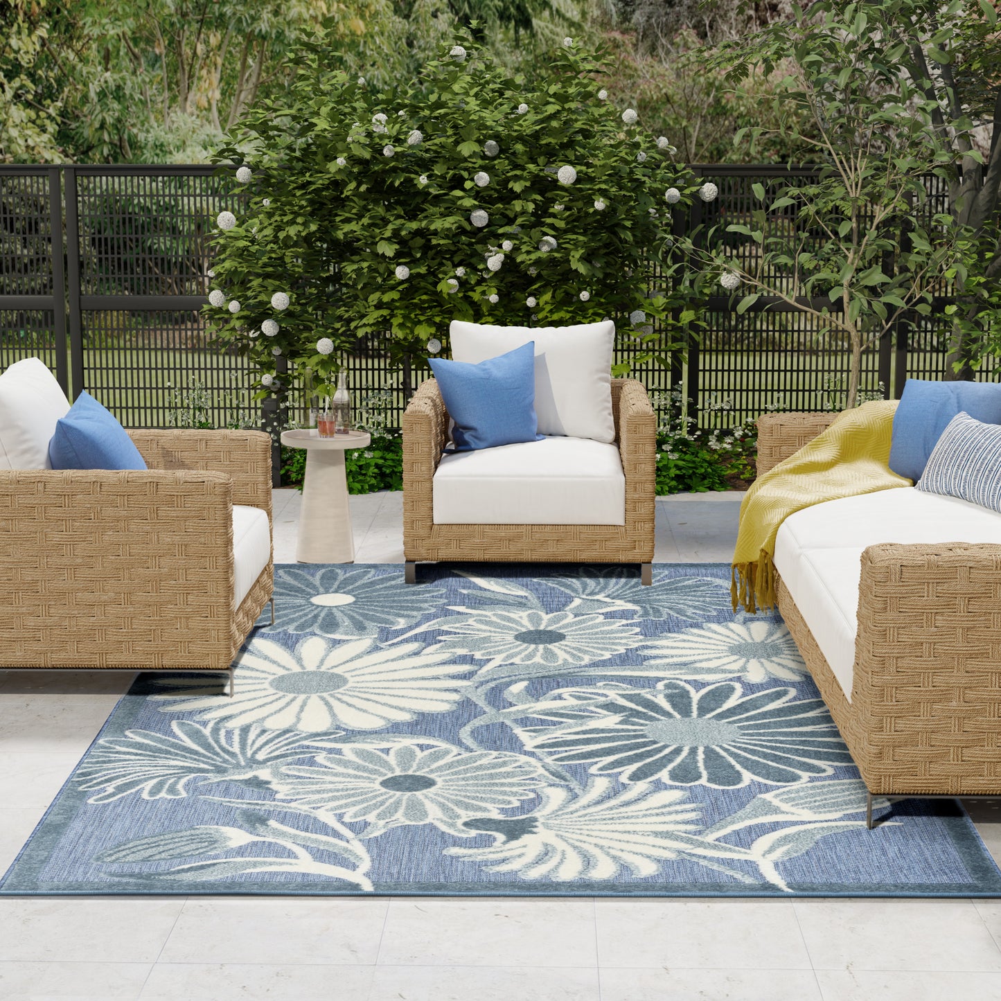 Outdoor wicker seating with white cushions on a blue floral rug, paired with blue pillows and a yellow throw.