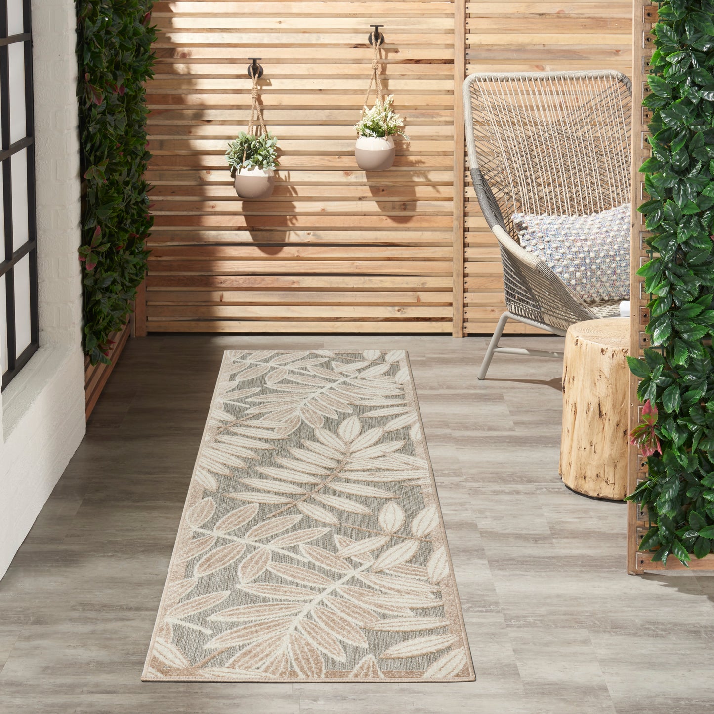Outdoor patio with beige leaf-pattern tropical 8' runner rug on gray wood floor, wicker chair, stump table, hanging planters, wooden slat wall.
