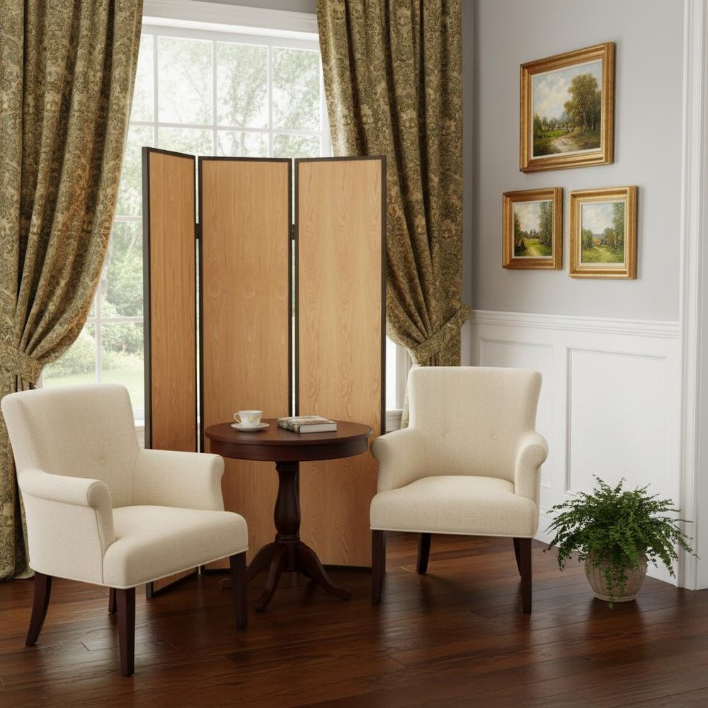 Two beige armchairs sit beside a round wooden table; folding screen, gold drapes, framed landscape art, and a plant.