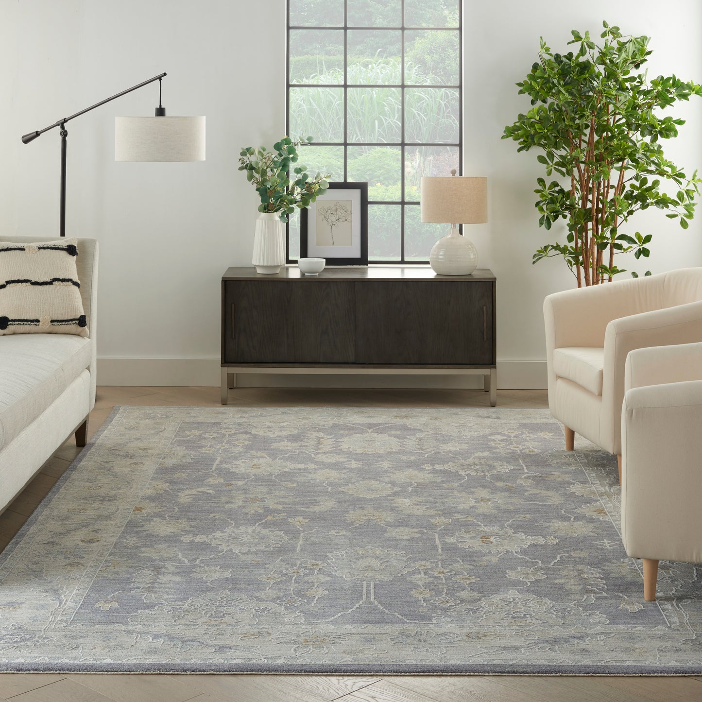 Neutral living room with dark wood sideboard, two cream lamps, gray-blue rug, beige sofa, armchair, and tall plant by window.