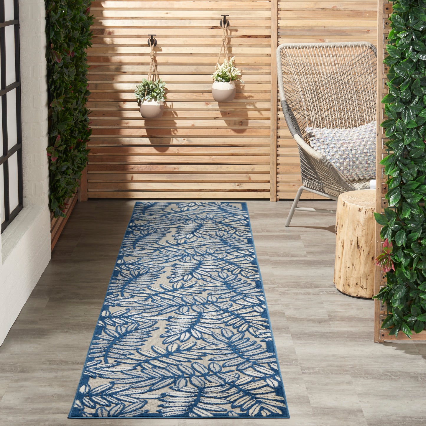 Outdoor rug runner with blue botanical leaf pattern on beige, laid on wooden deck beside wicker chair and hanging plants.