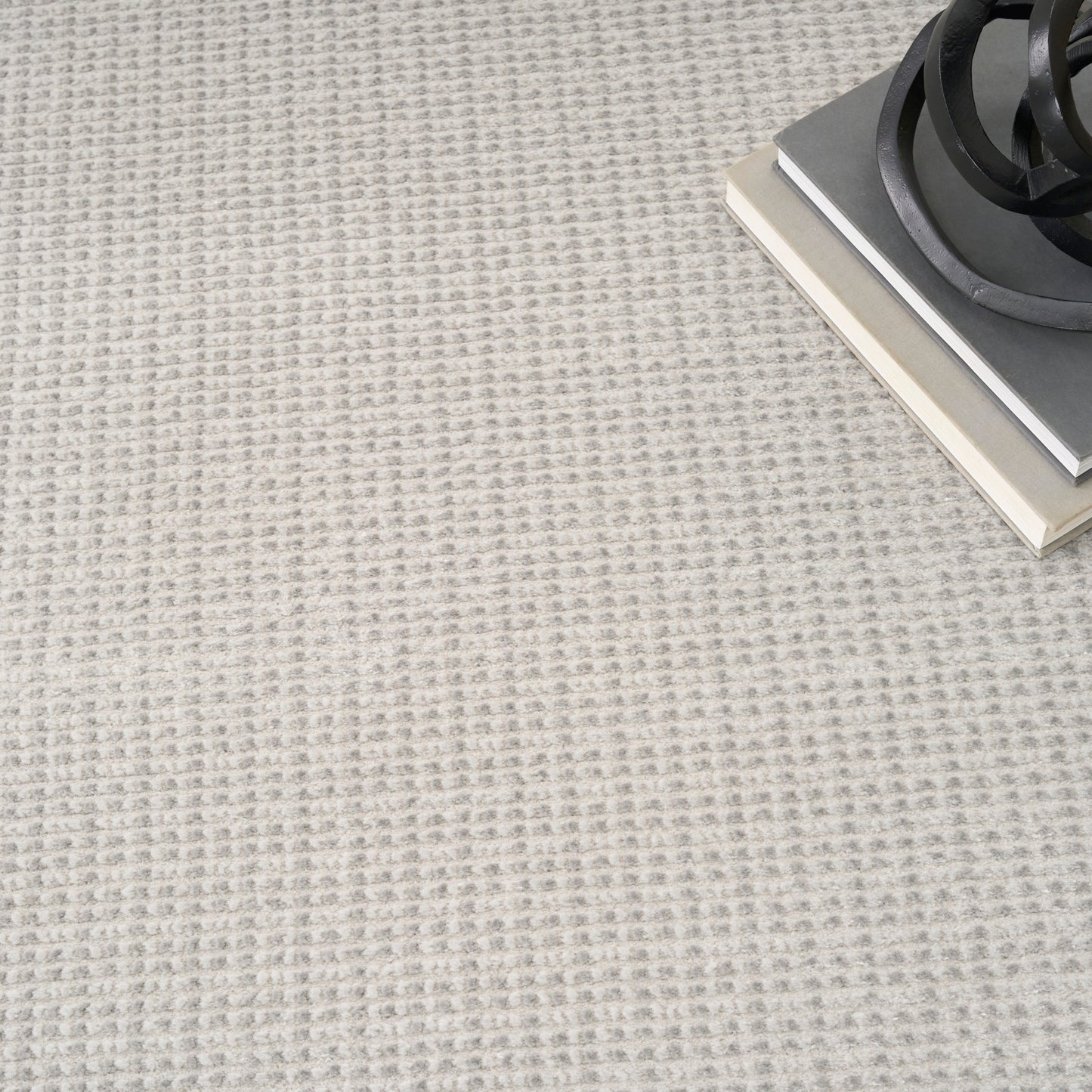 Beige textured rug with subtle square pattern; top-right shows a stack of gray books and a black spiral metal sculpture.