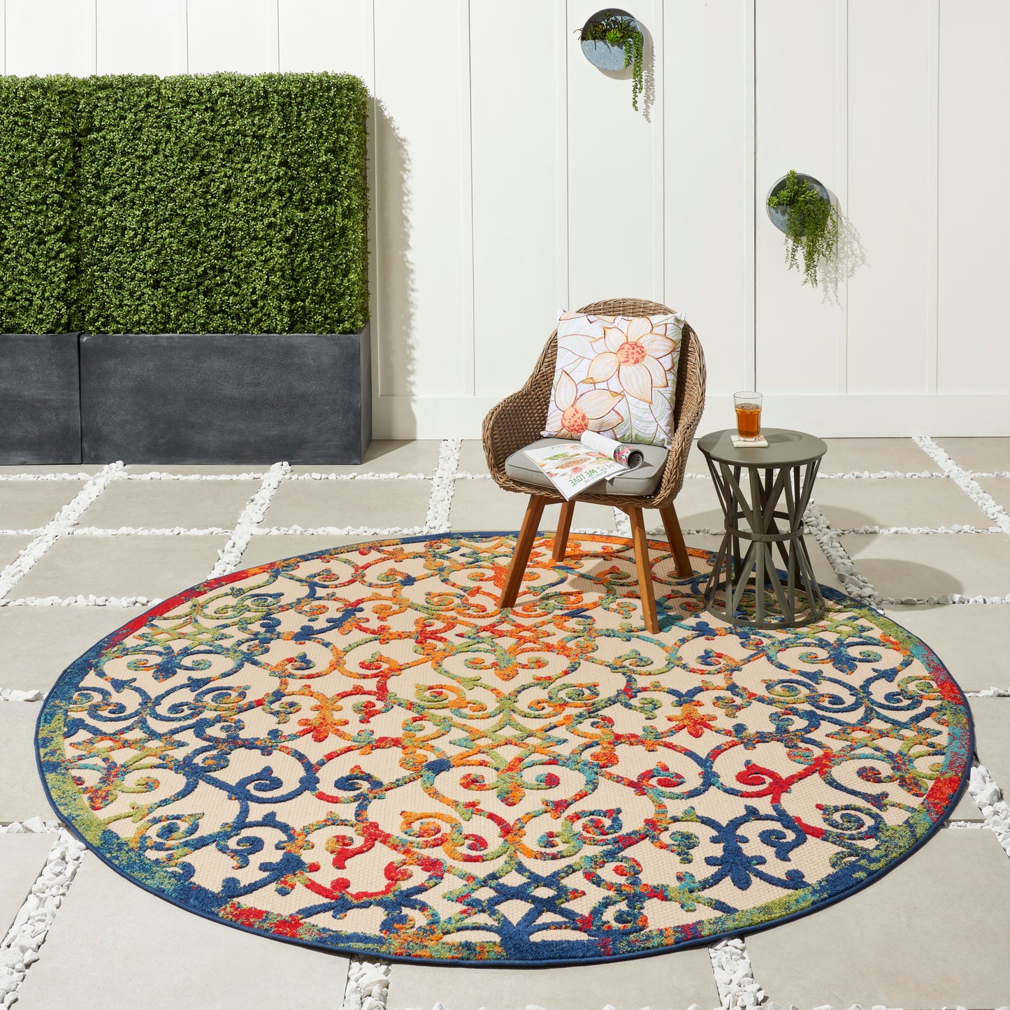 Round multicolor ornate rug on a patio with wicker chair, side table, cushion, glass of drink, white wall and greenery.