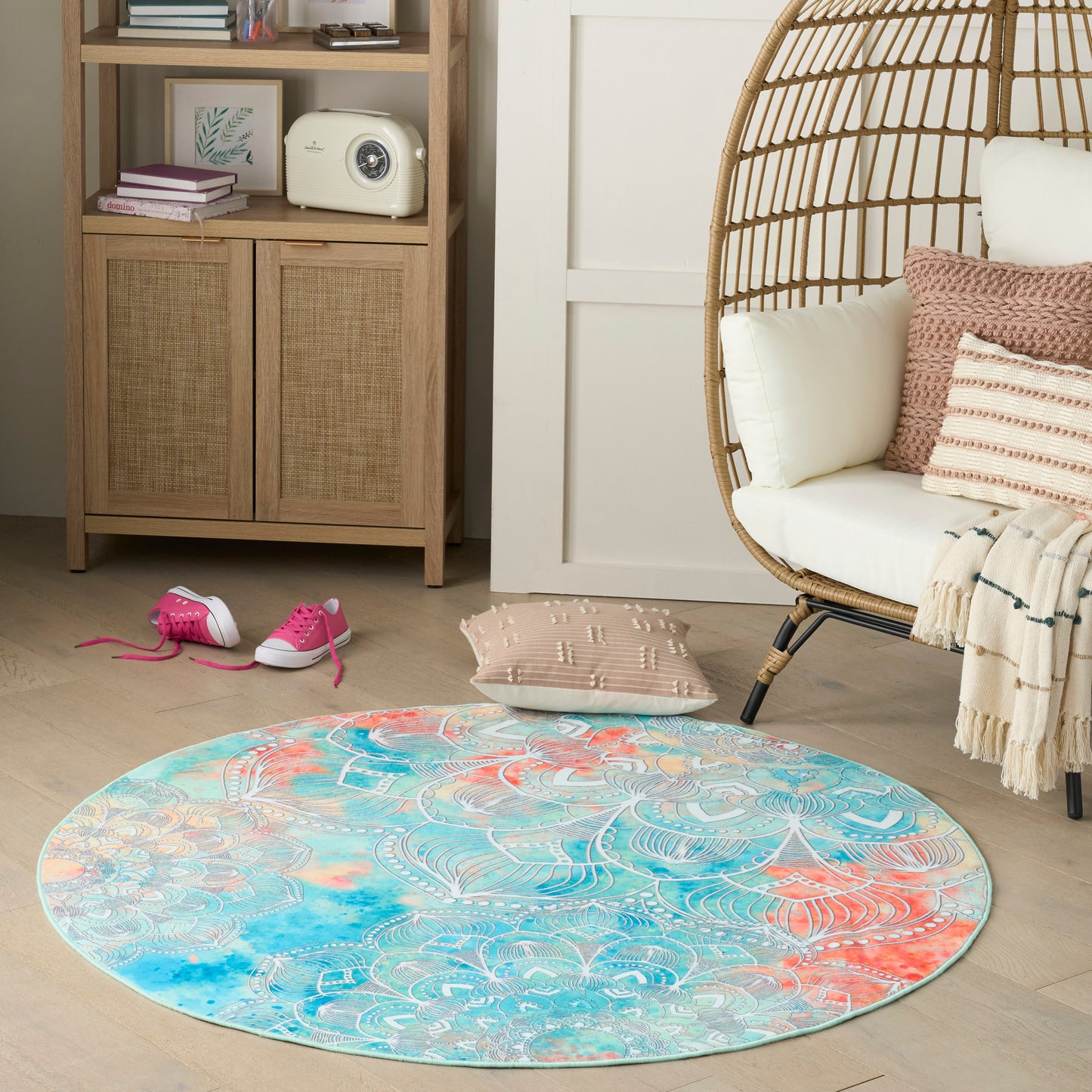 Round boho rug in turquoise, coral and teal with a white pattern, on light wood floor beside a wicker egg chair.