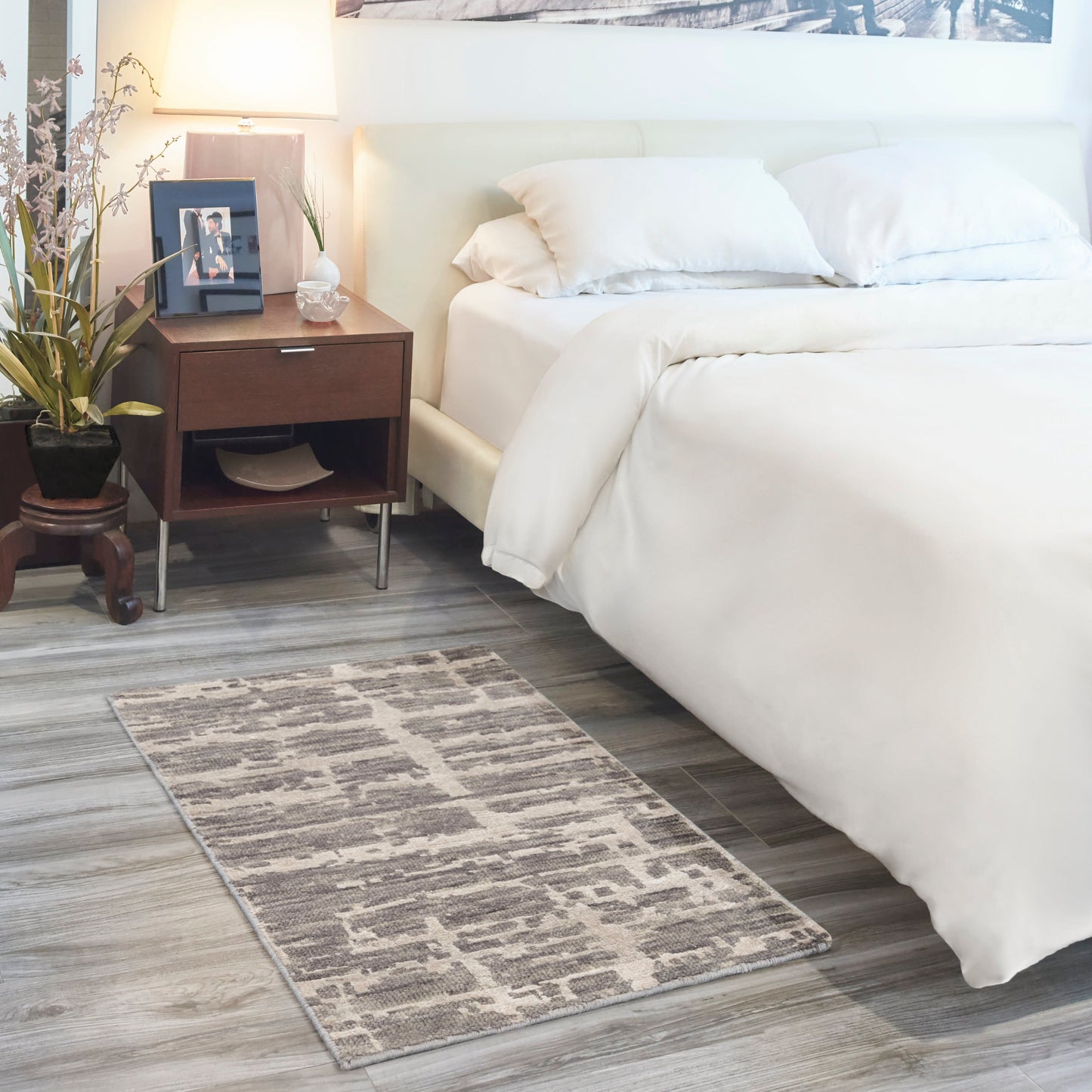 White upholstered bed with plush bedding, dark wood nightstand, lamp, framed photo, plant, and grey geometric rug.