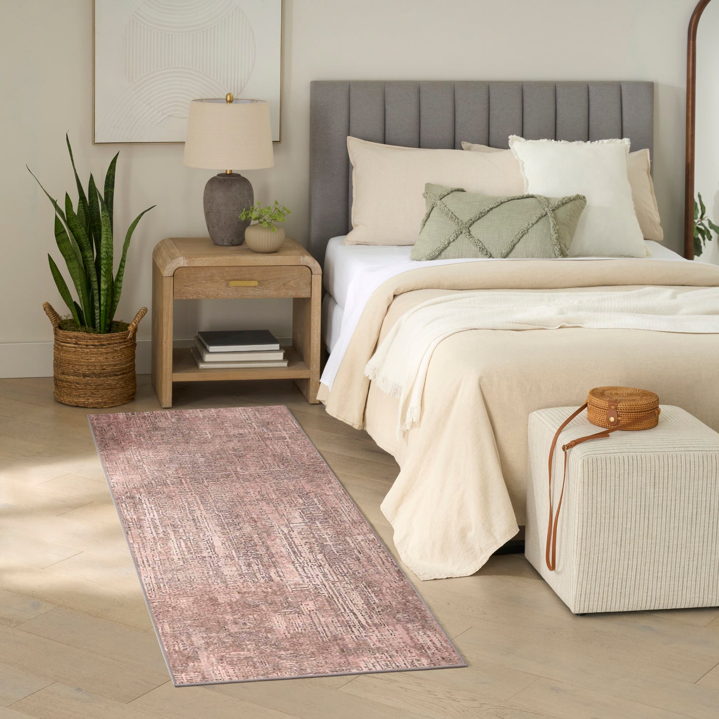 Neutral bedroom with tufted gray headboard, cream bedding, light wood nightstand, pink runner rug, potted plant, wicker basket.