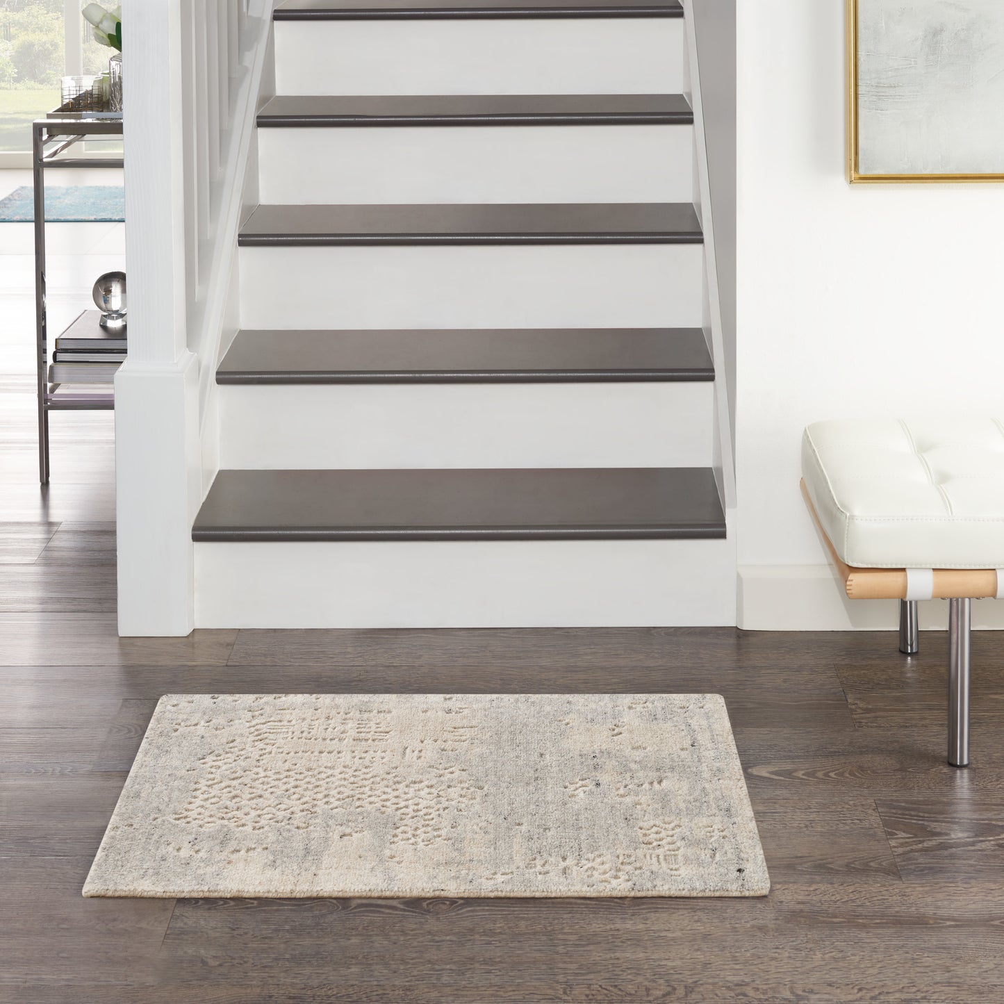Modern entryway with white staircase, dark brown treads, beige textured rug, white bench with chrome legs, & wall art.