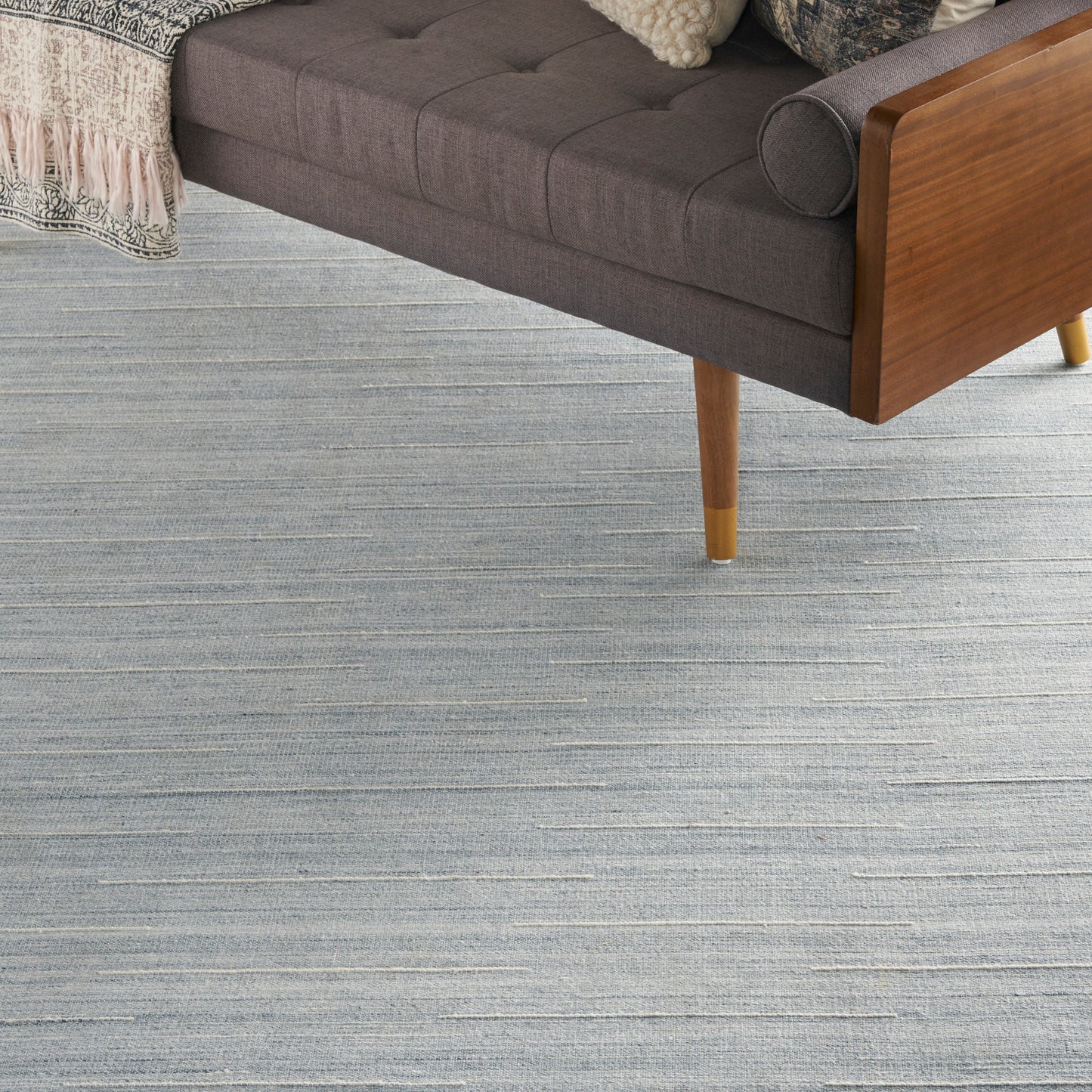 Dark gray mid-century sofa with wooden frame and tapered legs on a blue-gray striped rug; knit blanket draped.