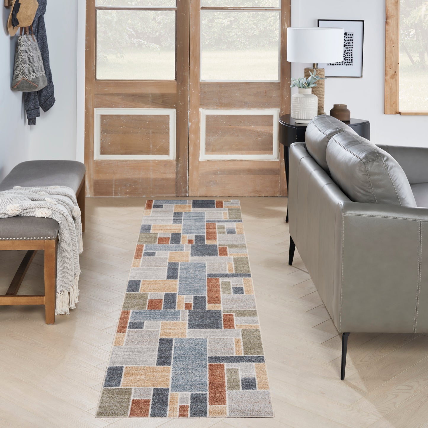 Long patchwork runner rug in blues, grays, beiges & rust; runs along light wood floor beside a gray sofa.
