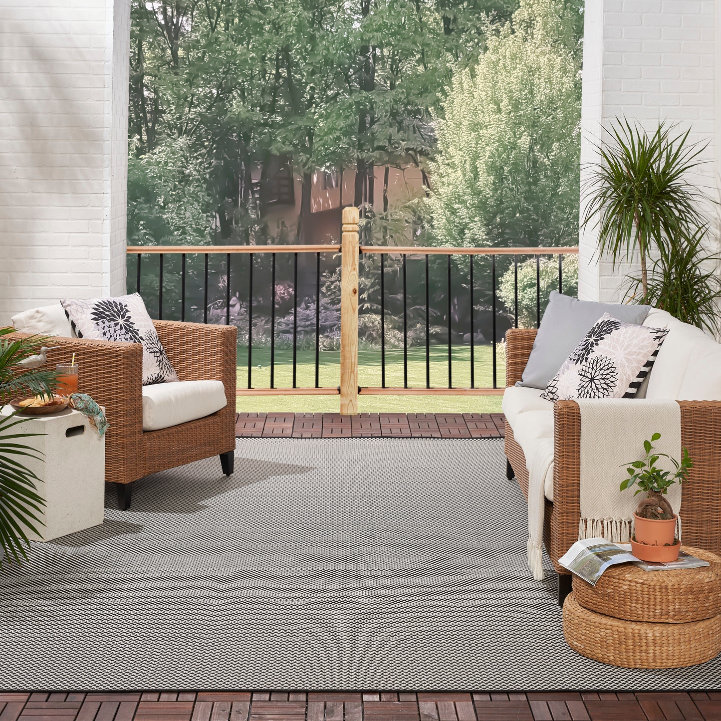 Gray rug on a brick porch under outdoor wicker seating with white cushions, patterned pillows, a round ottoman, potted plants, and a garden view.
