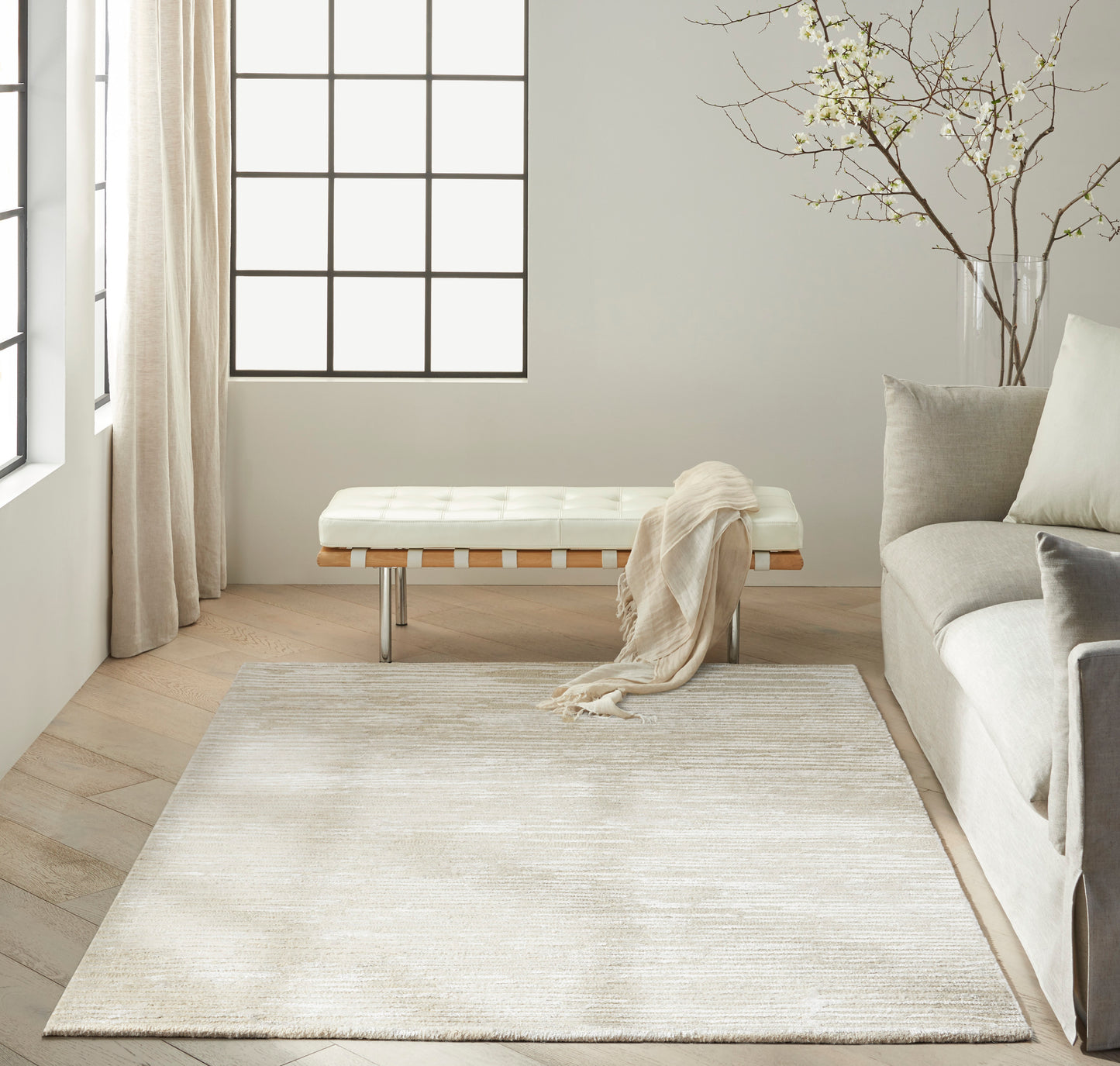 Sunny minimalist living room with a large pale beige rug, white tufted bench, soft throw, gray sofa, and grid windows.