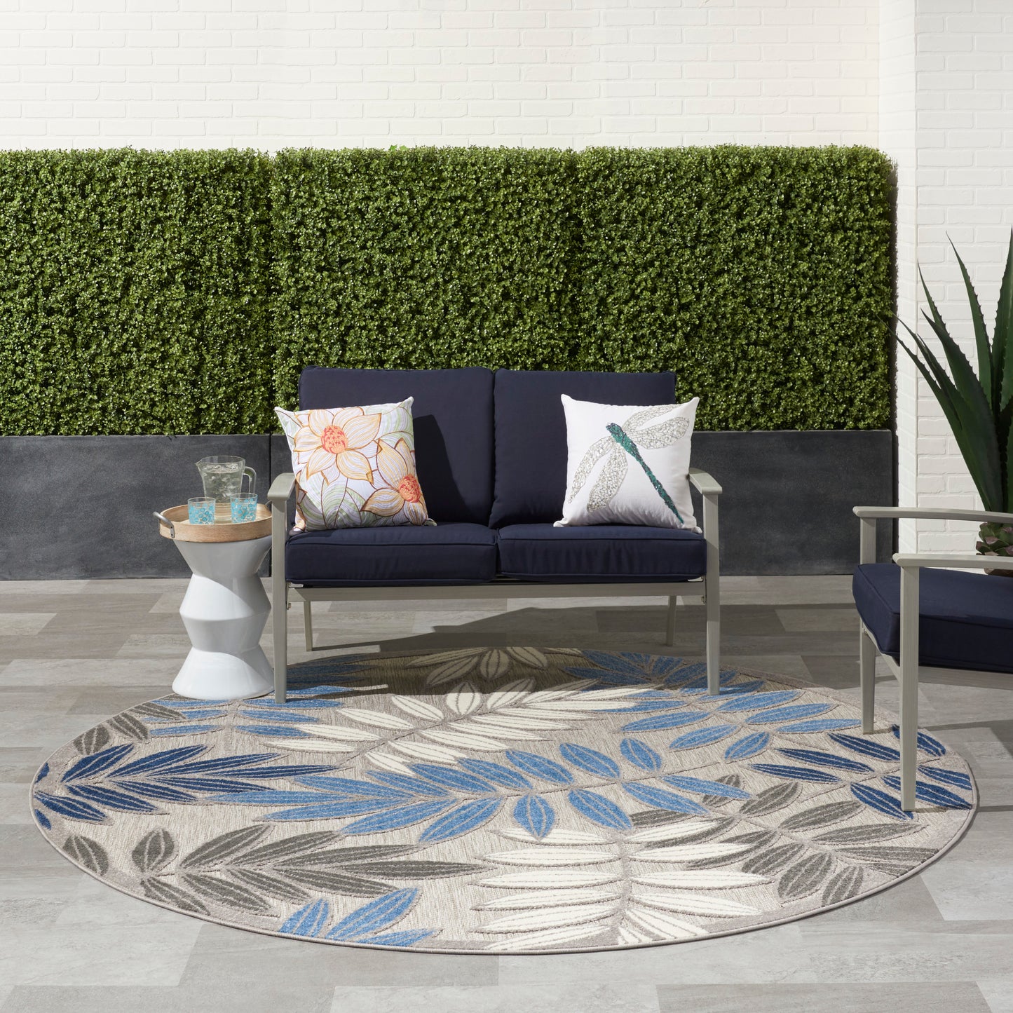 Round beige outdoor rug with blue and gray leaf pattern under navy cushioned patio sofa, side table, hedge wall backdrop.