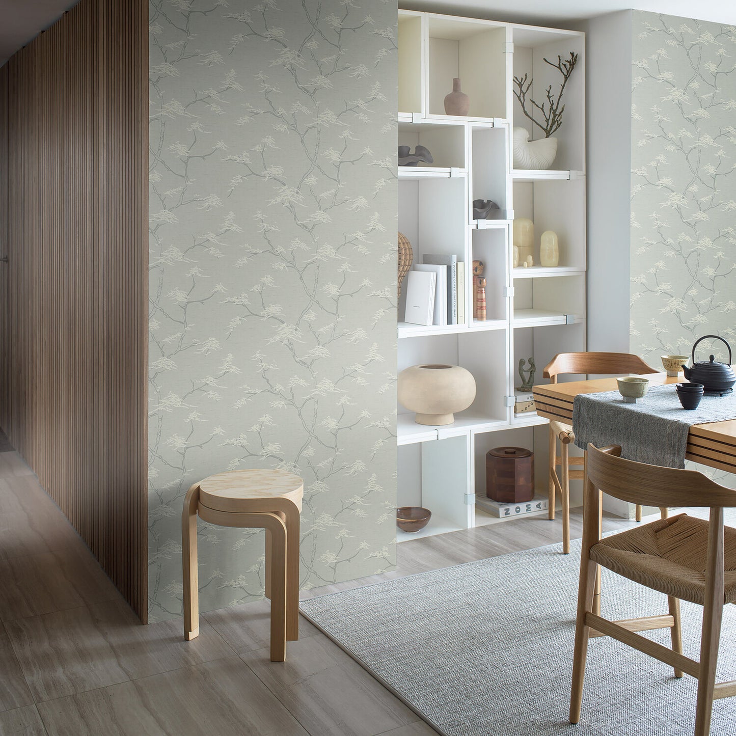 Soft green floral wallpaper, white open-shelf unit, light wood dining table and chairs, beige rug, wooden panel divider.