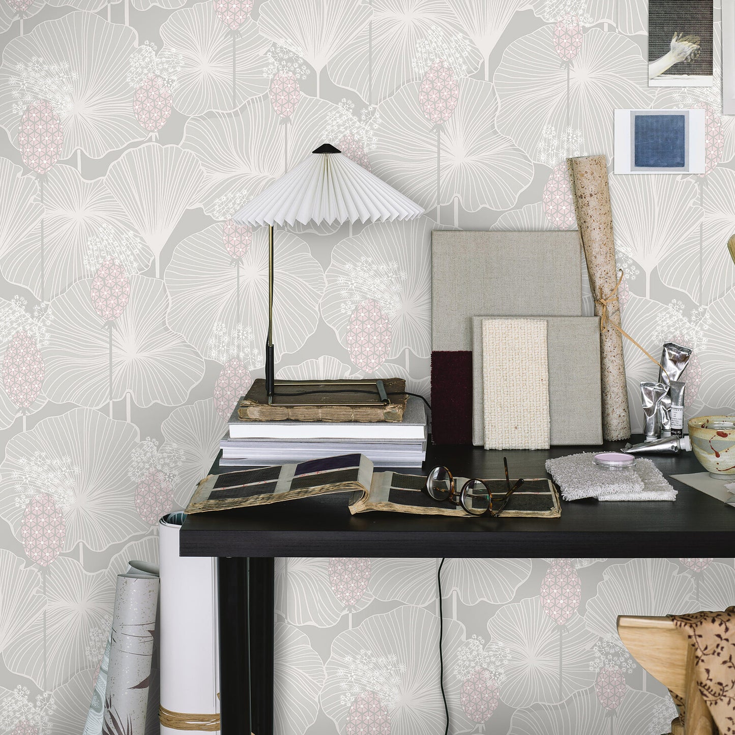 Black desk with fabric swatches, books, rolled canvases and a white pleated lamp, against pale floral wallpaper.