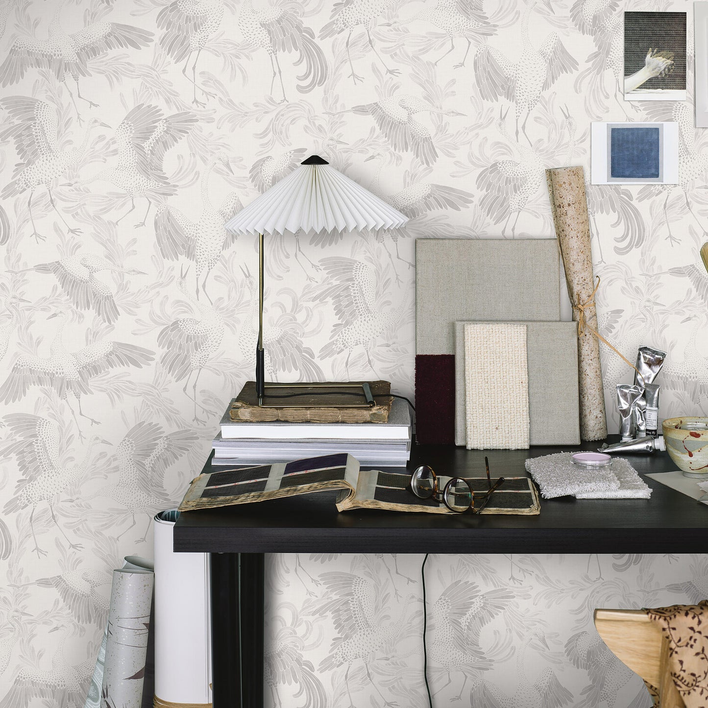 Black desk with fabric swatches, wallpaper rolls, and a pleated white lamp atop books against palm-pattern wallpaper.