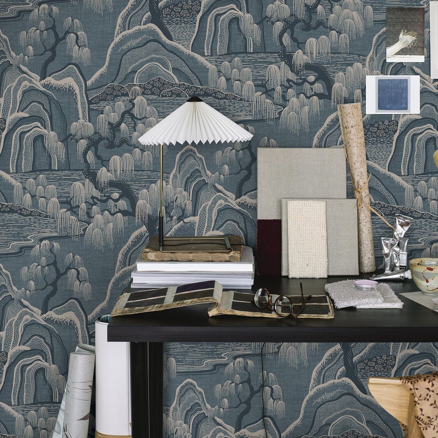 Blue-gray chinoiserie wallpaper backdrop; black desk with white pleated lamp, books, frames, rolled papers & glasses.