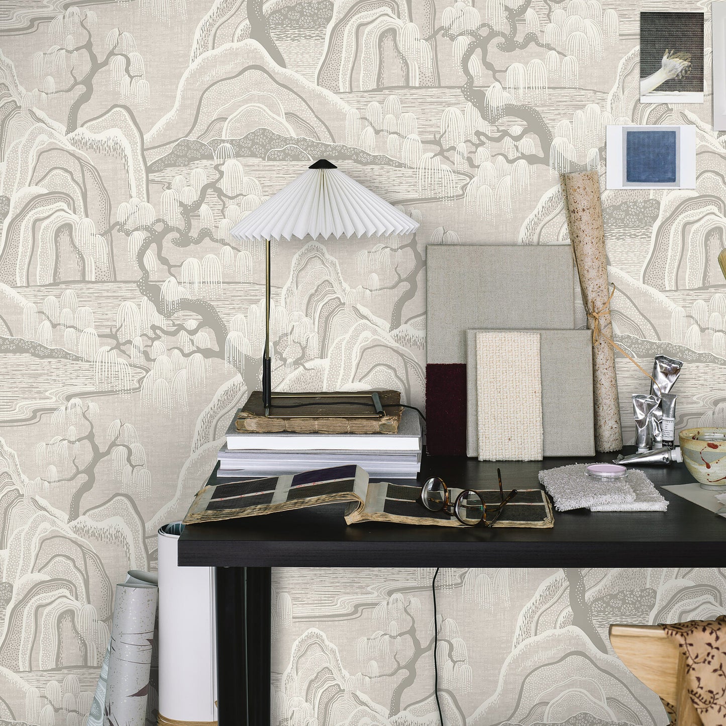 Dark wood desk with pleated white lamp, books, wallpaper samples, swatches and rolls against beige mountain-pattern wallpaper