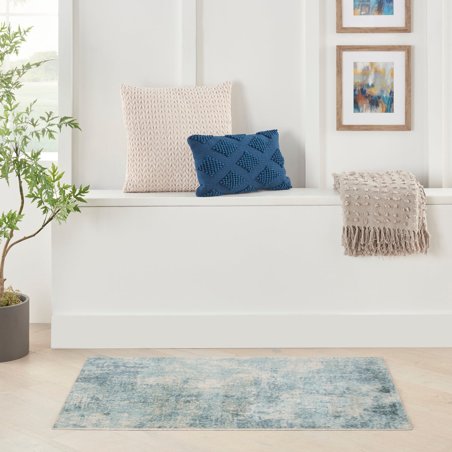 Beige knit pillow and beige fringed throw with a blue textured cushion on a white bench, plus plant and abstract art.