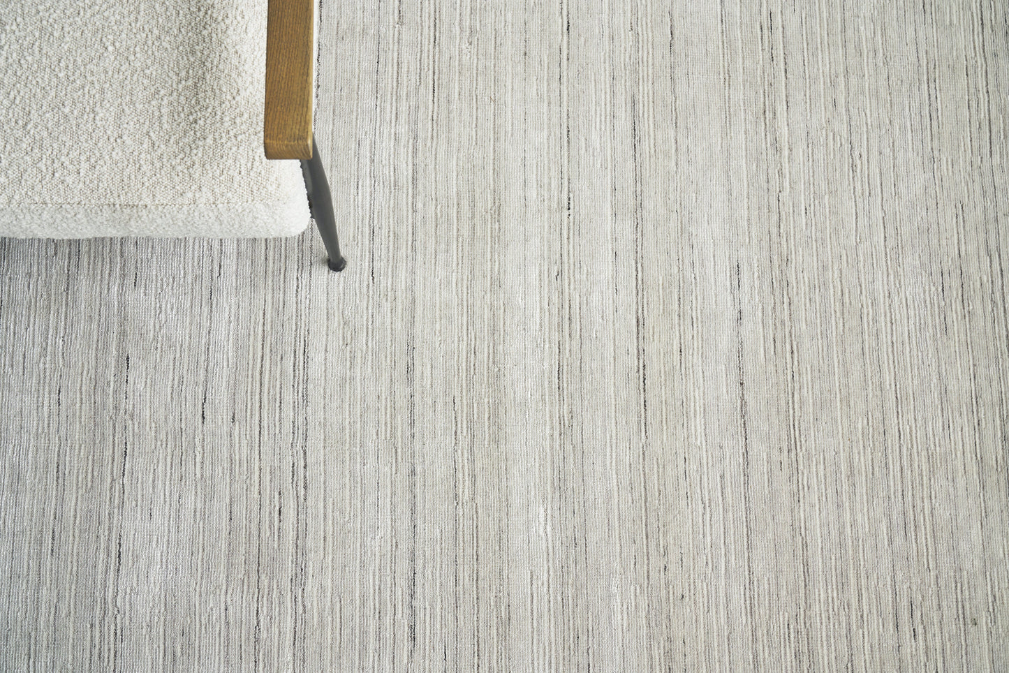 Close-up of a light gray textured rug with vertical stripes; corner of a chair with wooden arm & black metal leg.