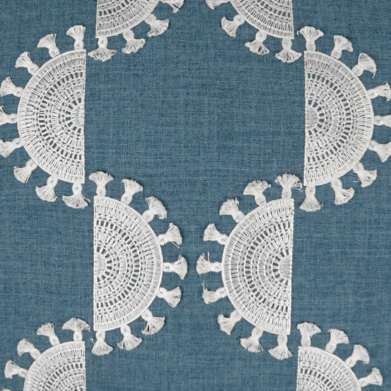 Blue woven fabric with white crochet doily-style circular motifs & tassel fringe; geometric repeating pattern.