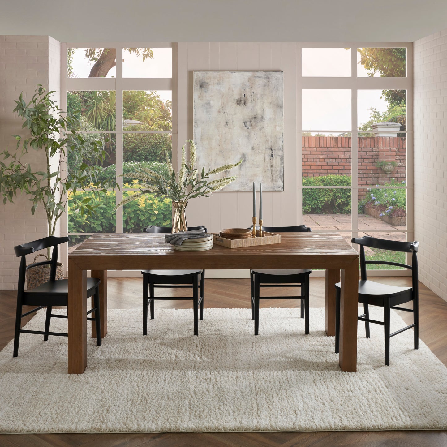 Cream shag rug under a rustic wooden dining table with black chairs, large windows with a garden view, abstract wall art.
